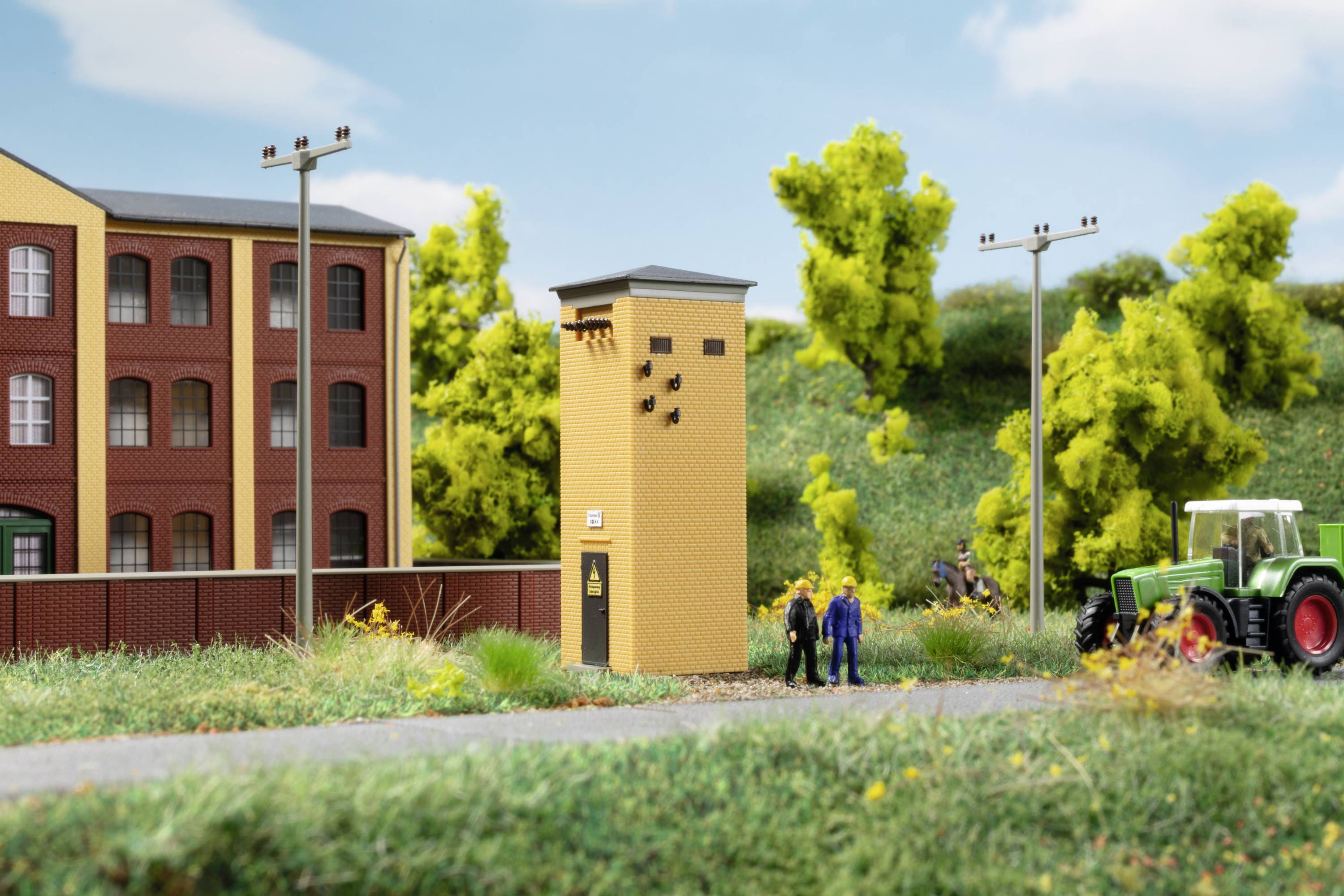 In einer ländlichen Umgebung steht ein Miniatur-Wasserturm neben zwei Figuren und einem Traktor, mit einer Backsteinfabrik im Hintergrund.