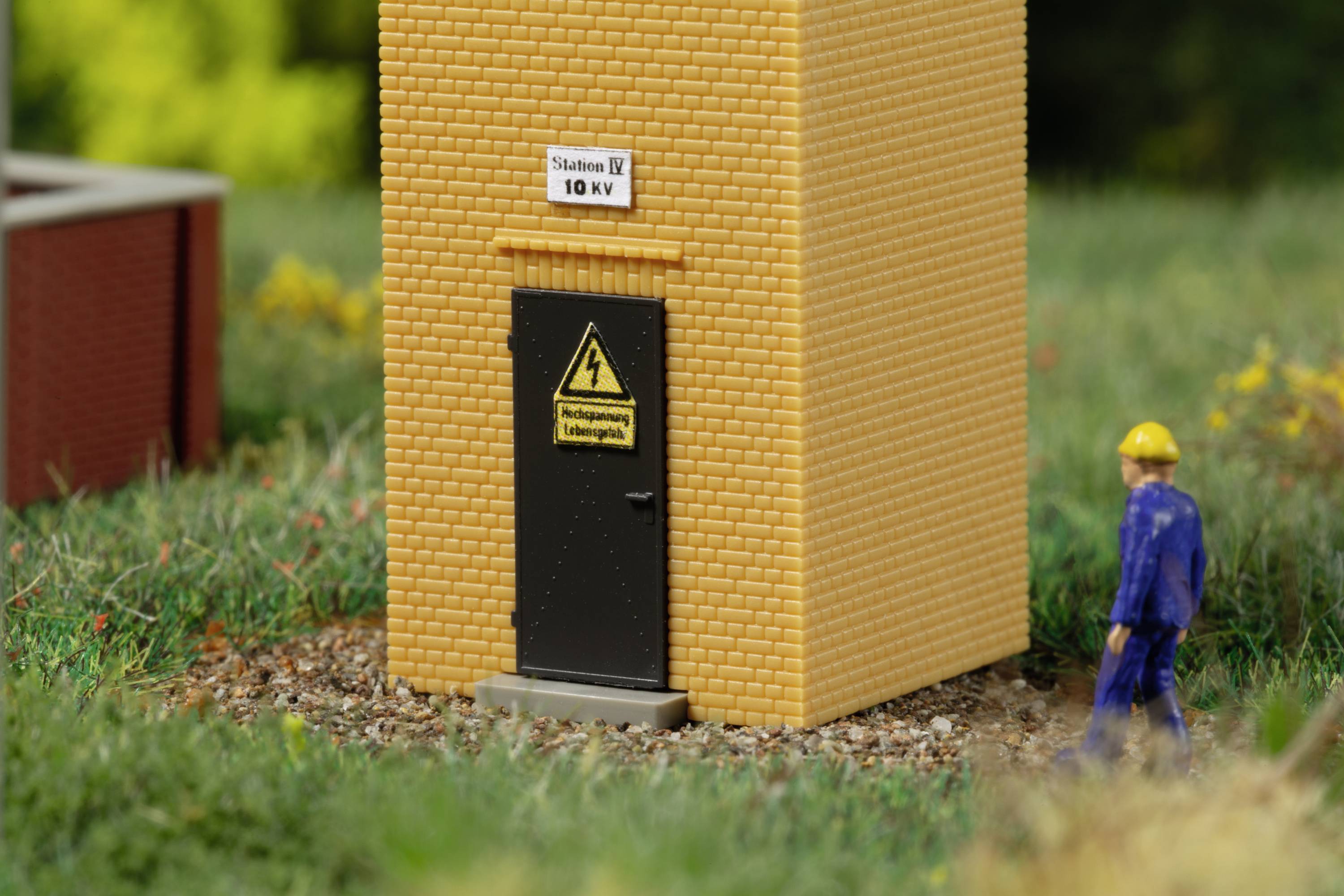 Ein Miniaturmodell zeigt ein gelbes Gebäude mit Tür und Warnschild 'Hochspannung Lebensgefahr'. Eine Figur in blauer Kleidung steht daneben.