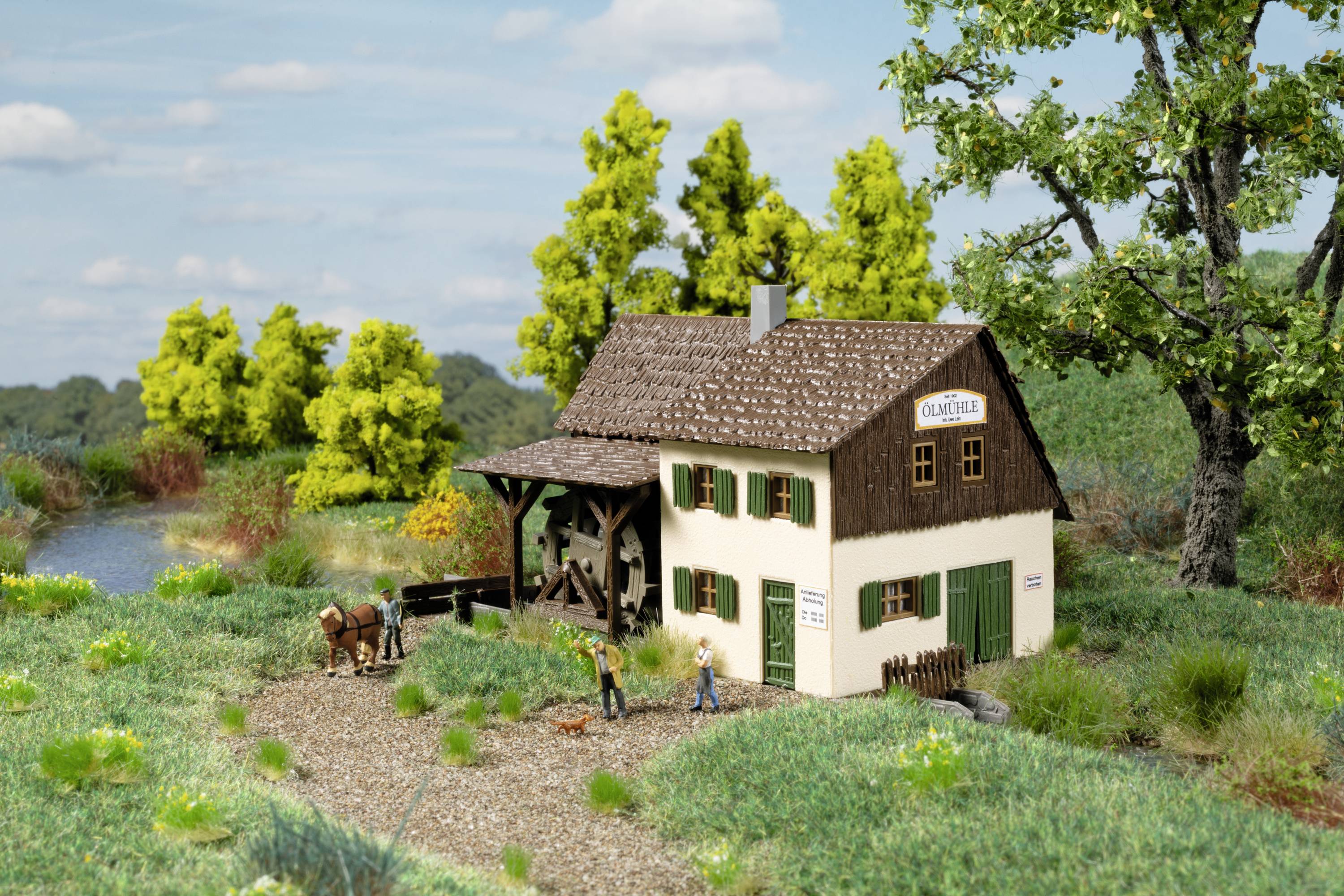 Ein Modellbau-Szenario zeigt ein Bauernhaus mit Strohdach und Holzveranda in einer ländlichen Umgebung. Zwei Figuren stehen vorne, ein Pferd ist in der Nähe.