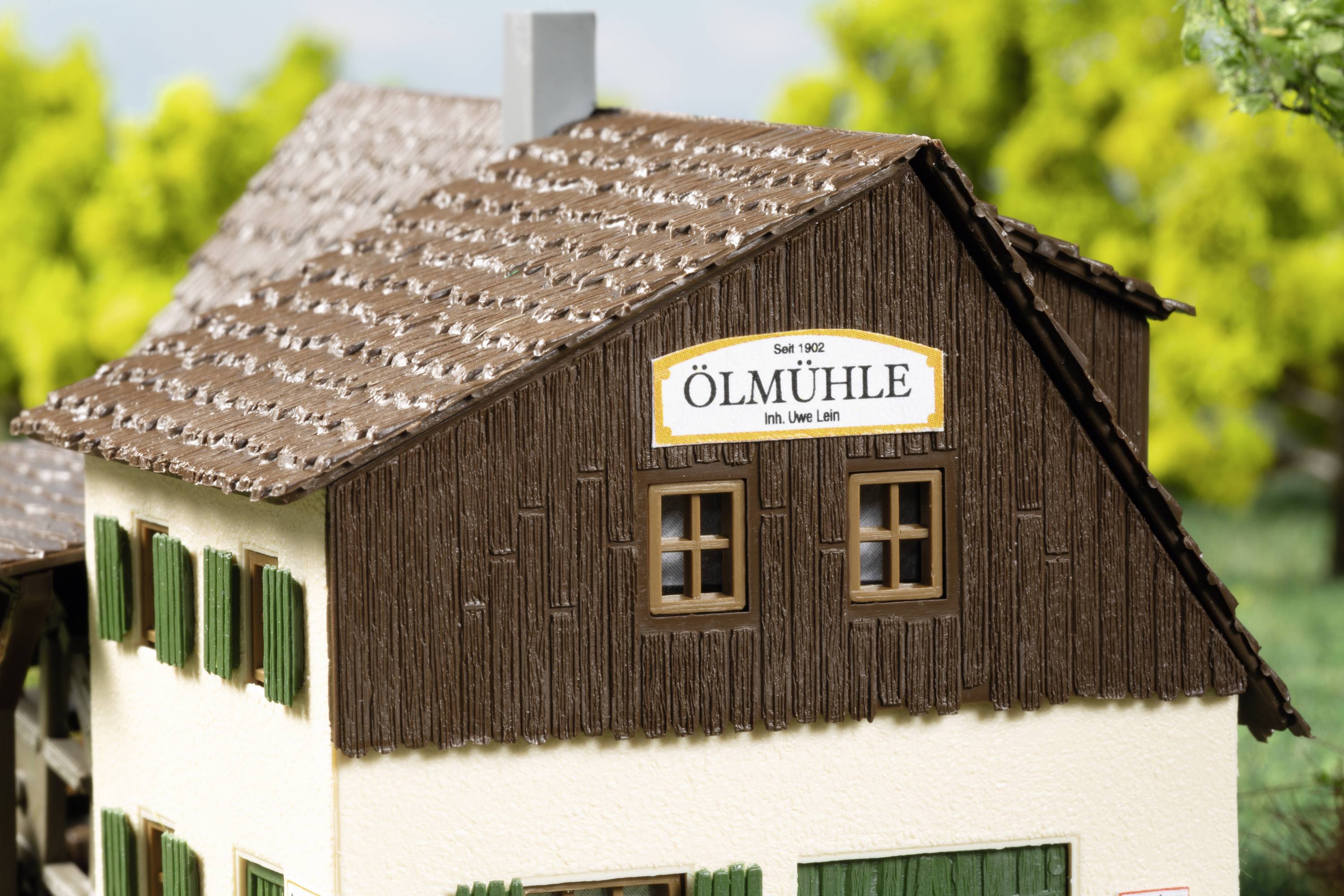 Ein Modell eines Hauses mit der Aufschrift 'Ölmühle' und grünen Fensterläden vor einem Hintergrund aus Bäumen.