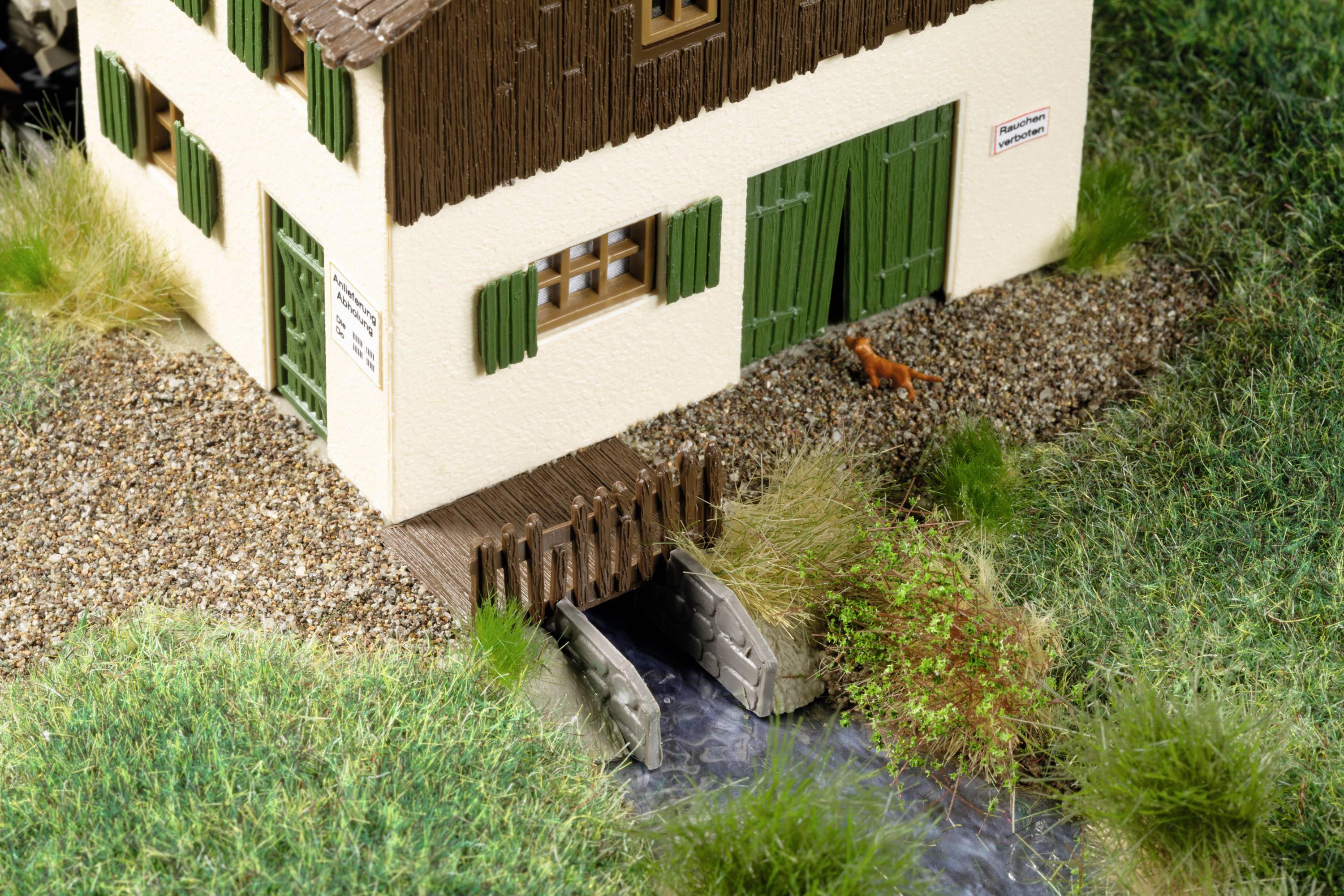 Modell eines zweistöckigen Gebäudes mit grün gestrichenen Holzläden und Türen, daneben ein kleiner Bach und eine miniaturisierte Katze.