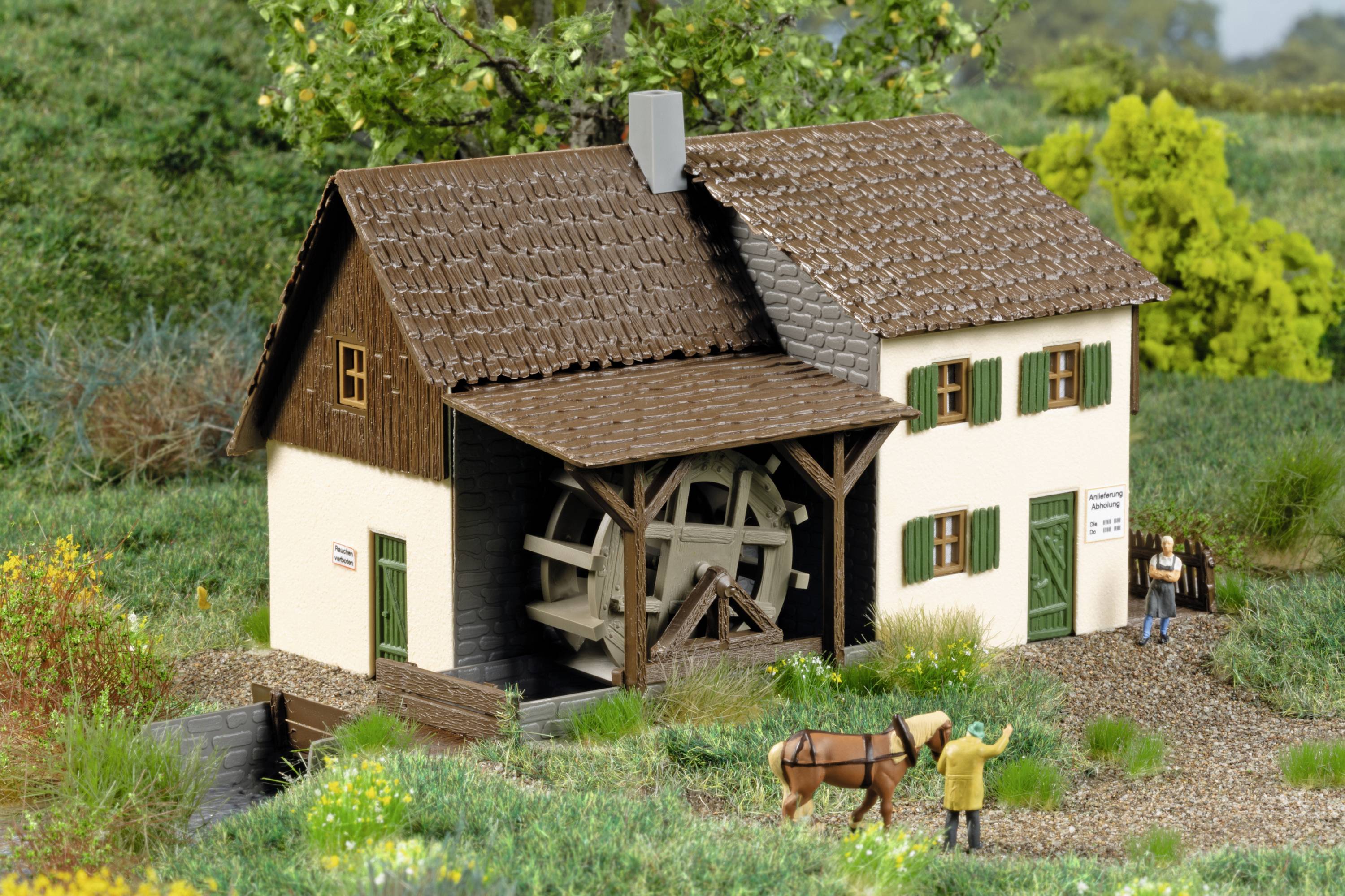 Miniaturmodell einer Wassermühle mit drehendem Mühlrad, umgeben von grüner Landschaft. Vor der Mühle steht eine Figur mit Pferd.