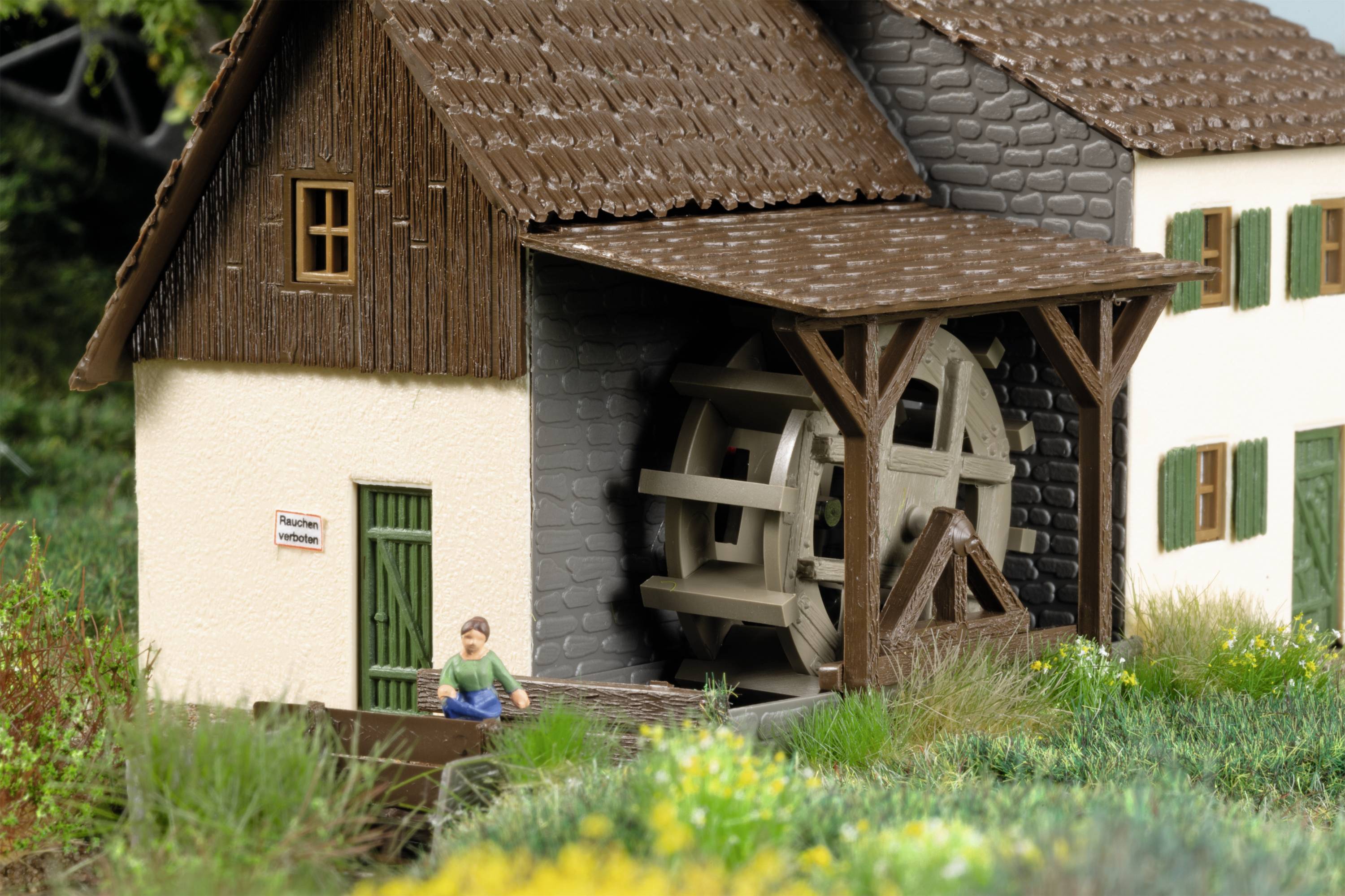 Modell eines Hauses mit Wasserrad. Vor dem Holzgebäude sitzt eine Miniaturfigur. Umgebung mit Gras und Blumen.