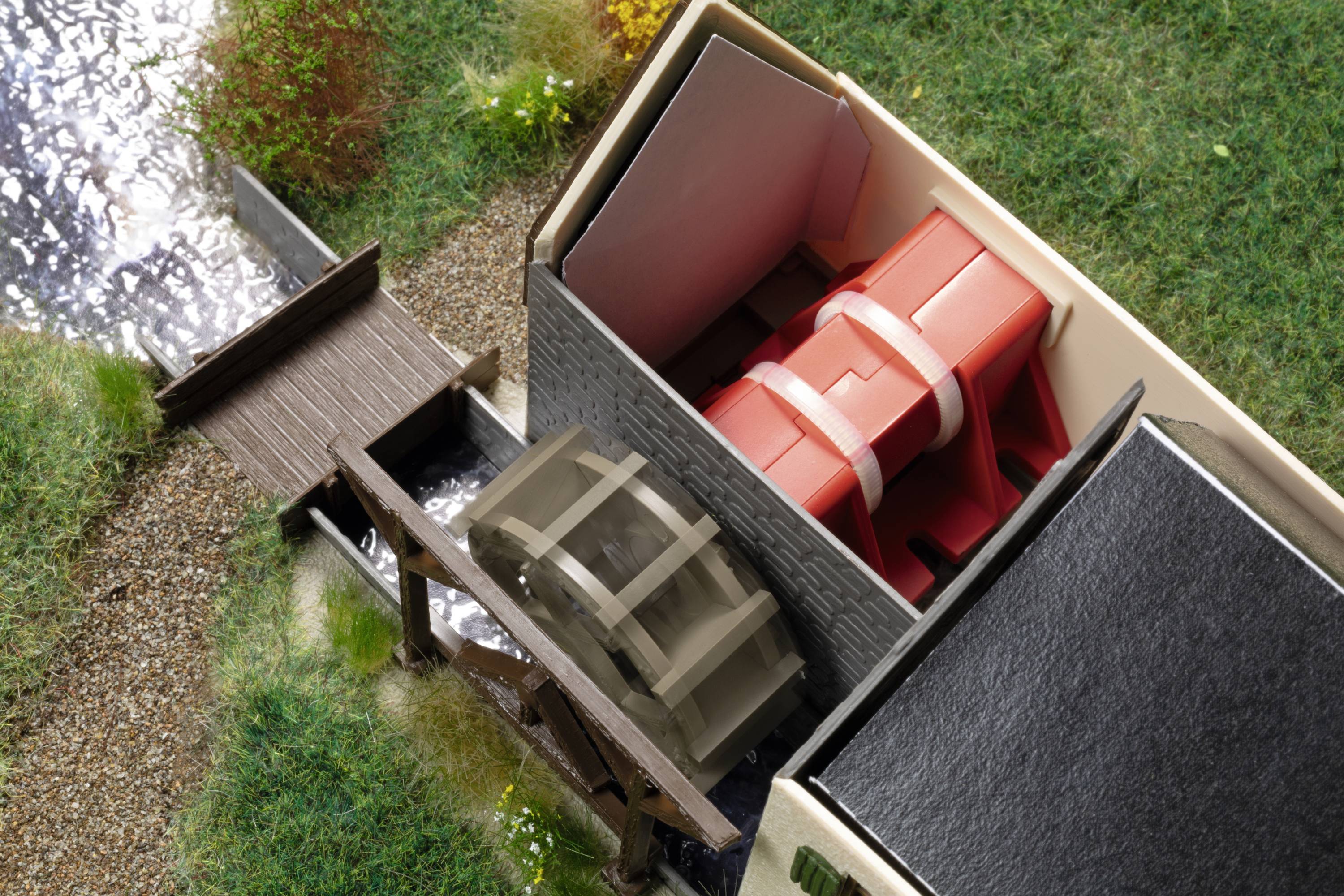 Eine Miniatur-Wassermühle zeigt ein großes, rotes Rad im Inneren eines Gebäudes mit einem kleinen Bachlauf und Holzbrücke daneben.