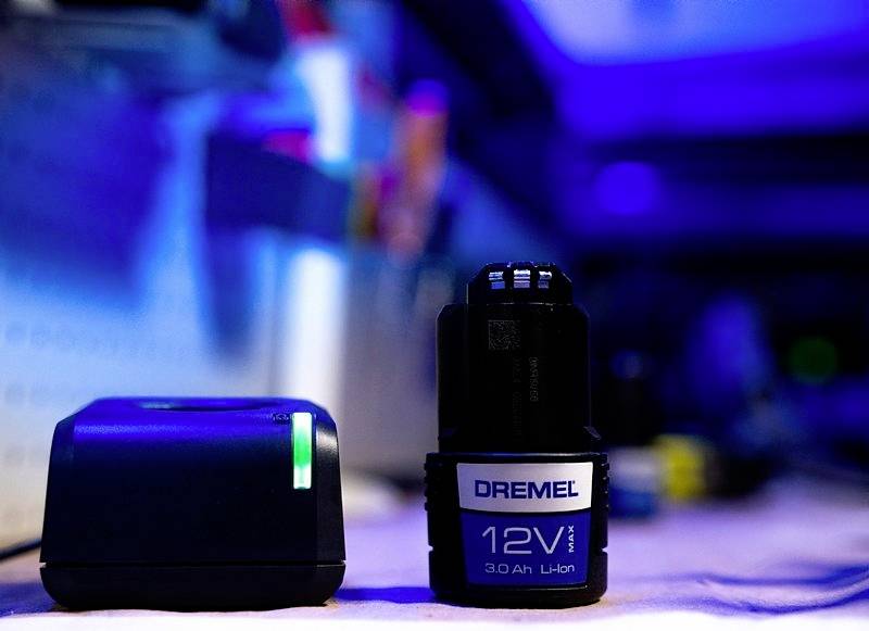 Ein 12V Dremel Lithium-Ionen-Akku und ein Ladegerät auf einem Tisch in einer Werkstattumgebung mit blauer Beleuchtung.