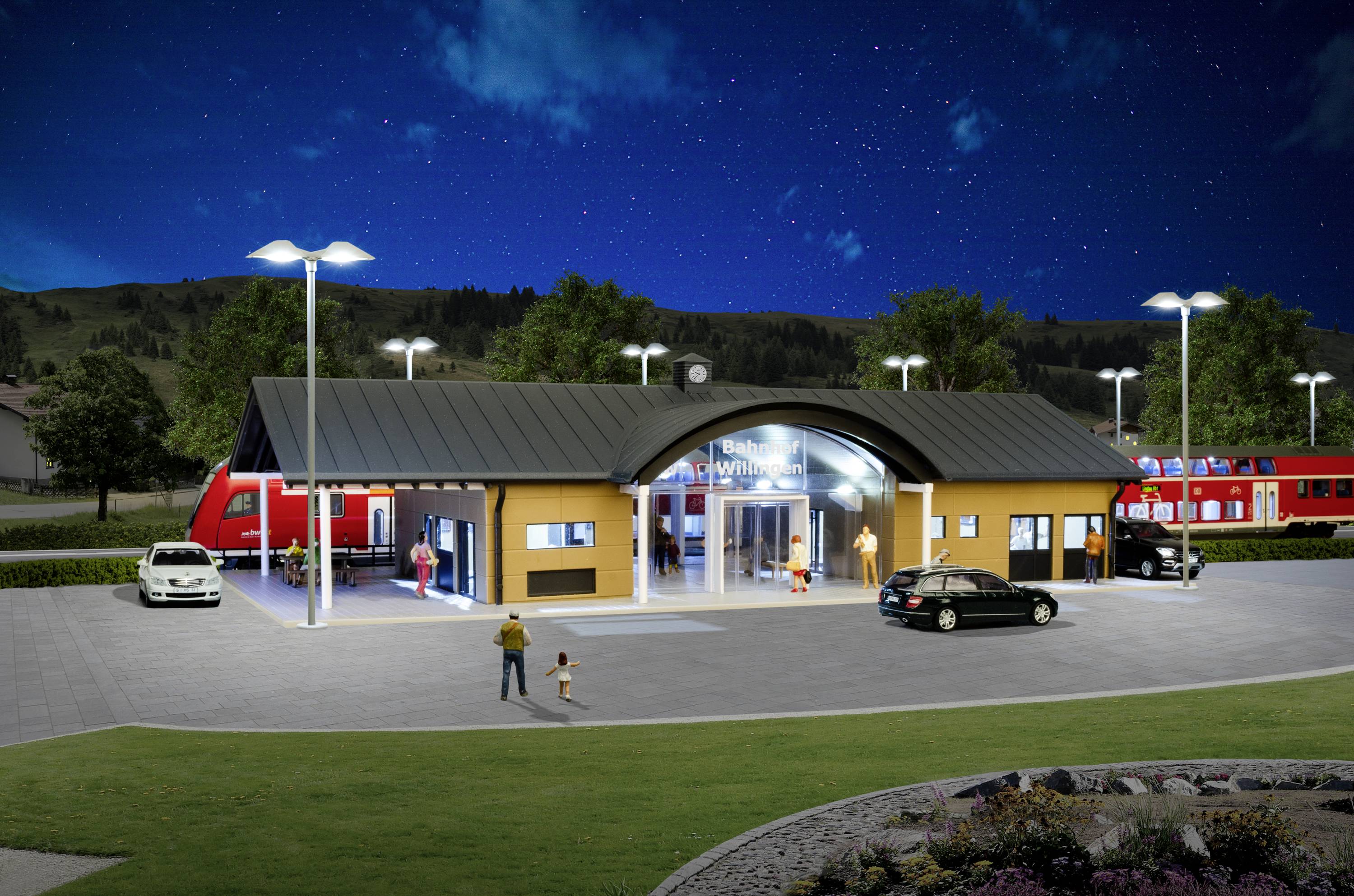 Ein kleiner Bahnhof bei Nacht mit mehreren geparkten Autos, Personen, die sich bewegen, und einem abfahrenden Zug auf einem Gleis im Hintergrund.