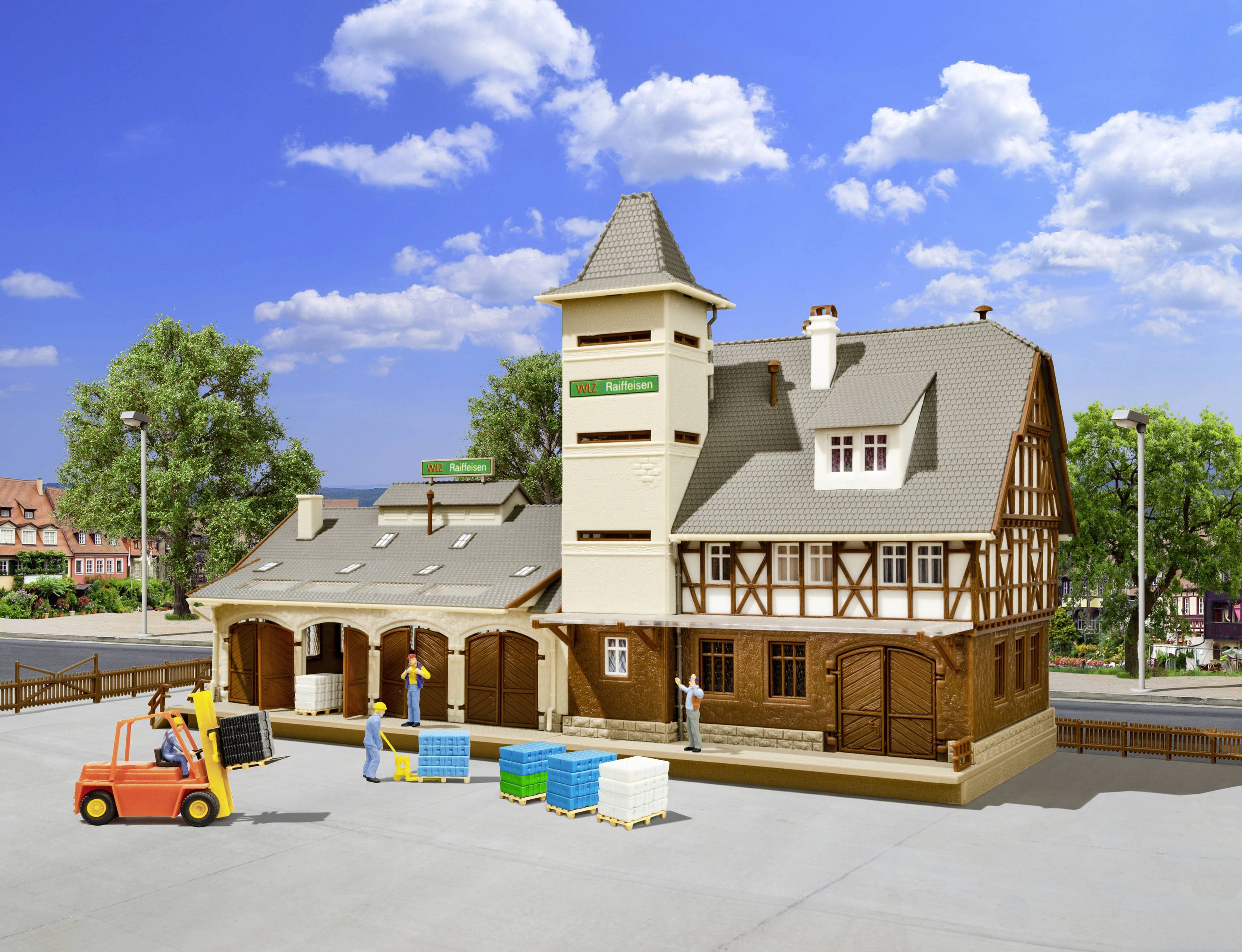 Spielzeugmodell eines Bahnhofs mit Güterlager, Gabelstapler und Figuren auf einer betonierten Fläche, umgeben von Bäumen und Himmel.