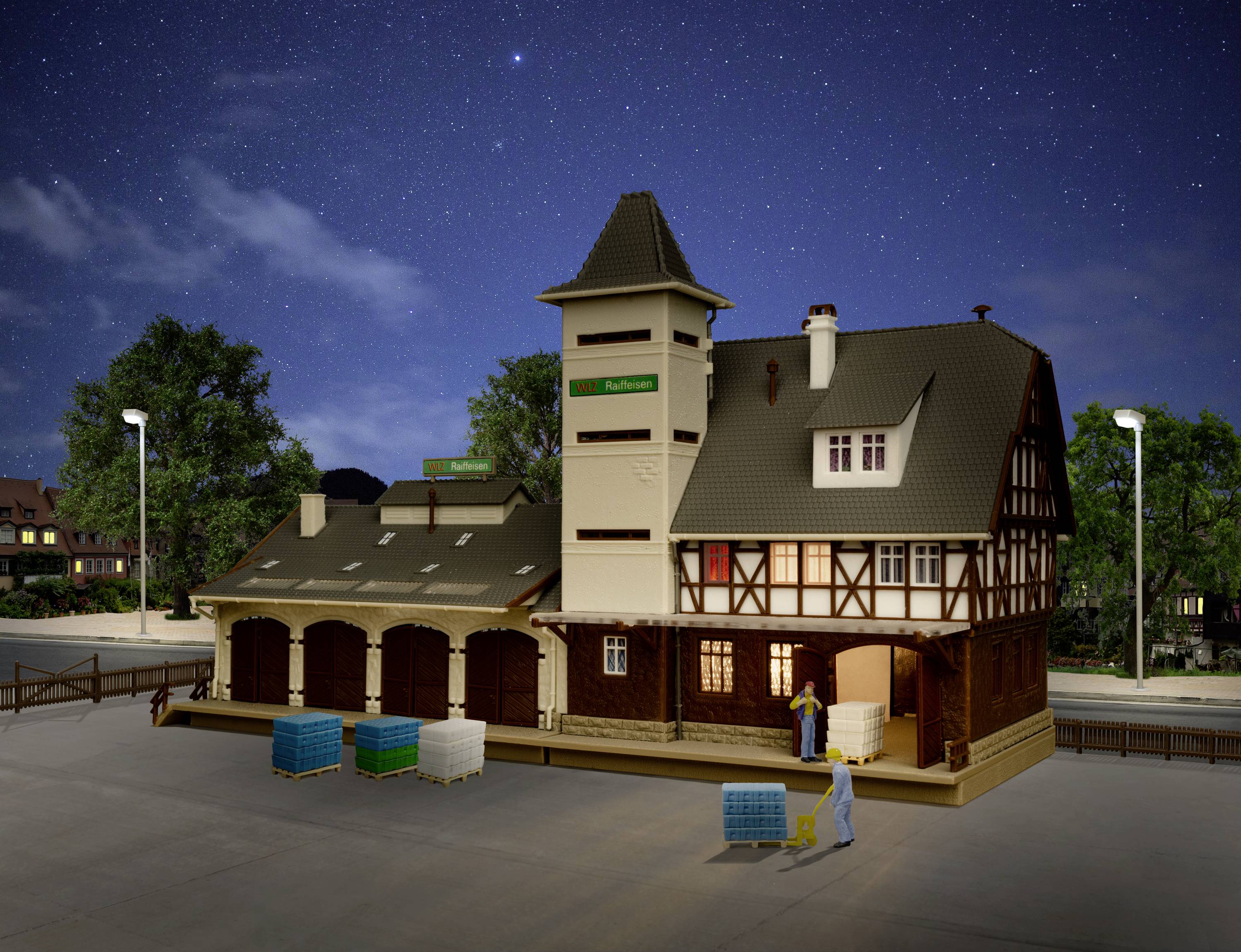 Ein Modellgebäude im Nachbau eines historischen Bahnhofs bei Nacht, beleuchtet von Straßenlaternen. Zwei Figuren beladen eine Plattform mit Gütern.