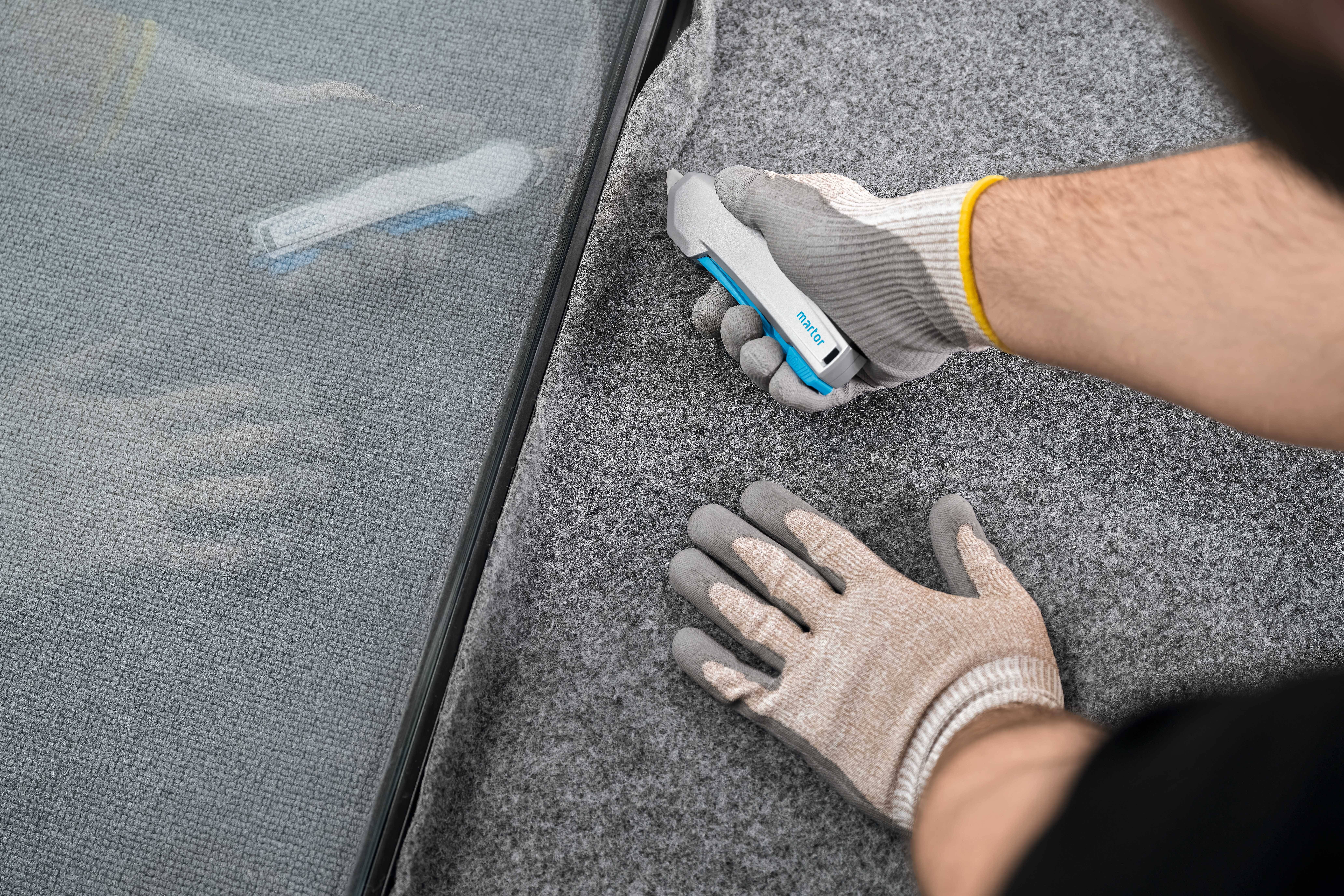 Person schneidet mit einem Teppichmesser grauen Teppichboden nahe einer Glaskante. Hände tragen Arbeitshandschuhe.