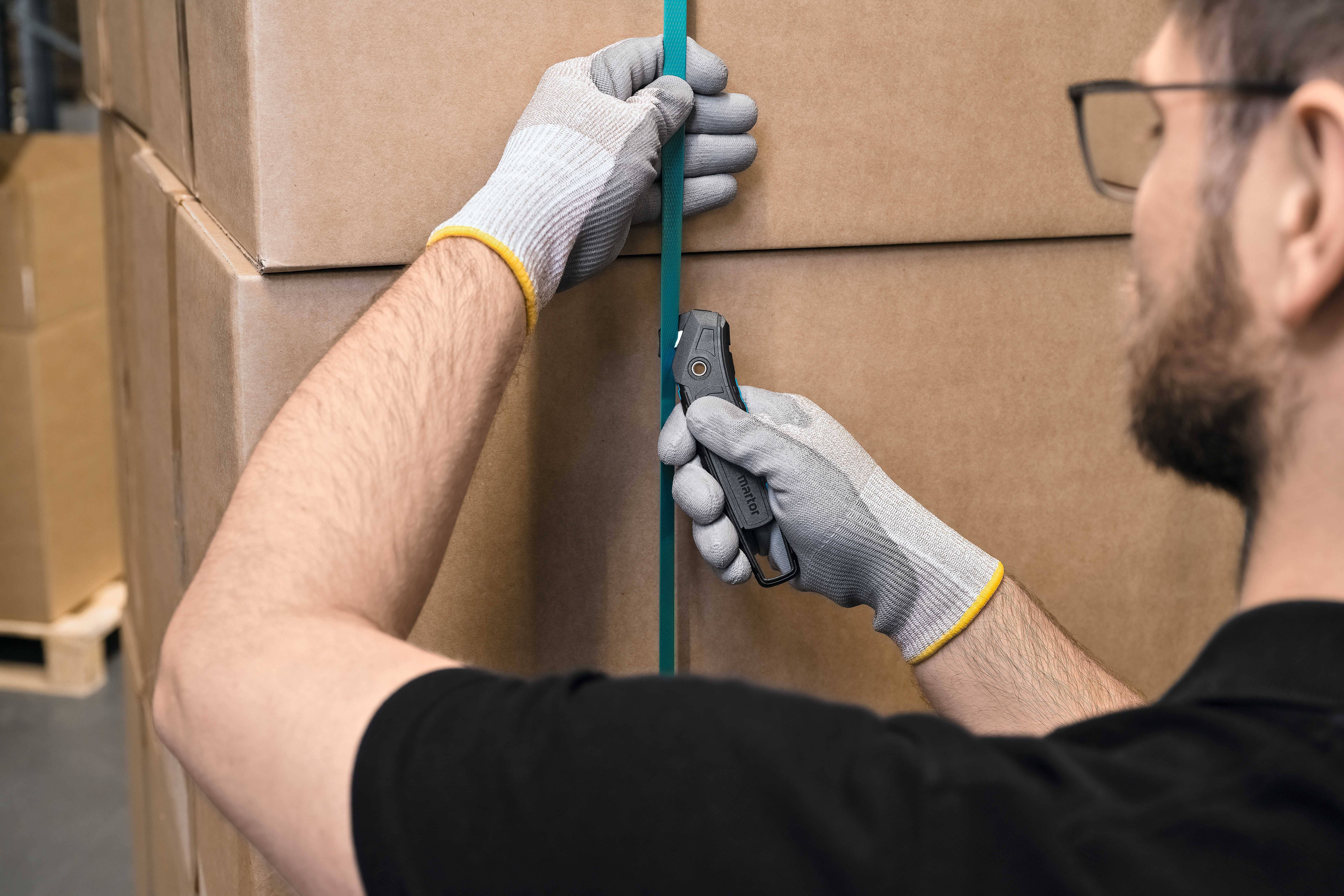 Eine Person in Handschuhen schneidet ein grünes Band von einem Kartonstapel mit einem Sicherheitsmesser in einem Lager.