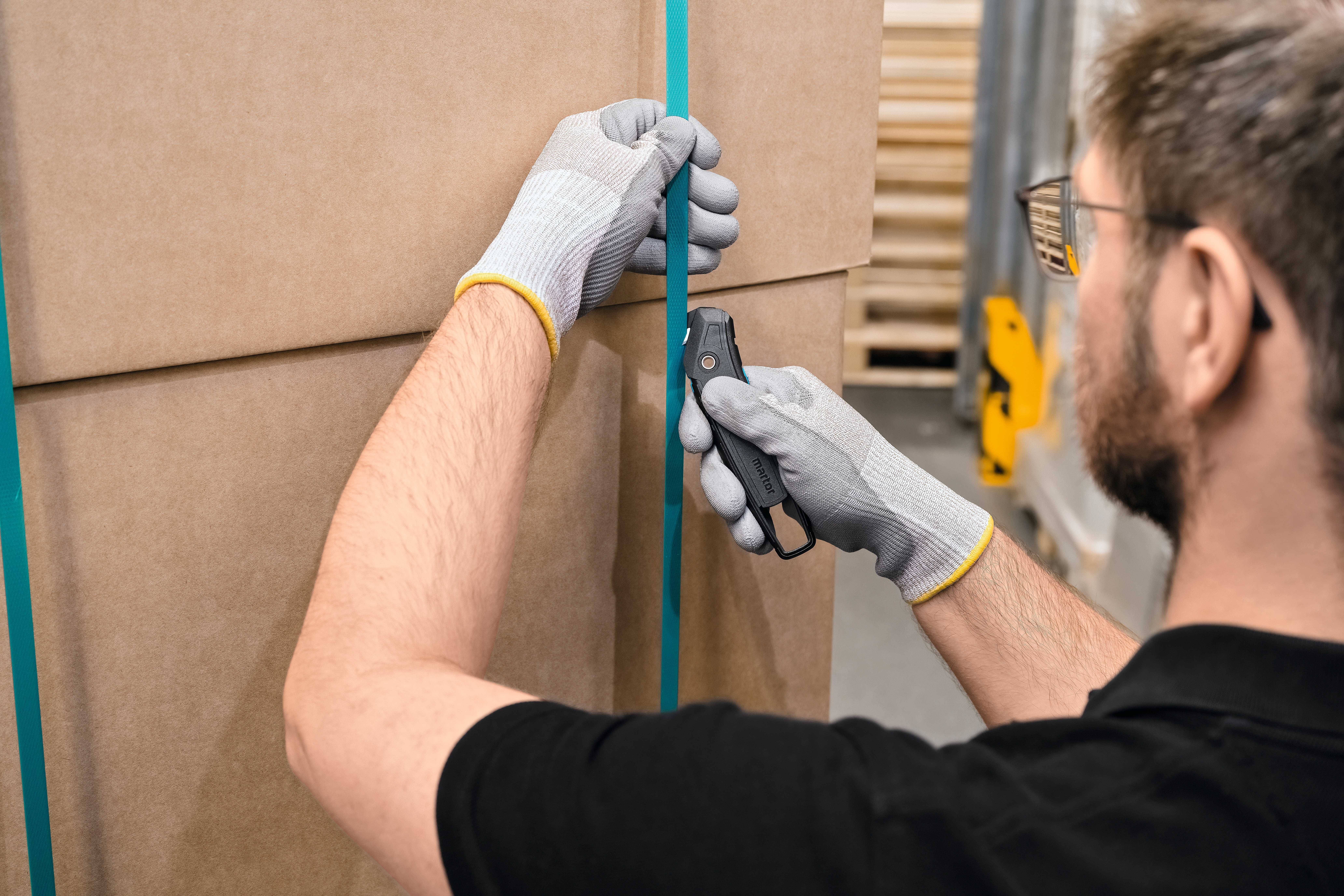 Mann mit Handschuhen öffnet Kartonverpackung mit einem Messer. Verpackung ist mit blauem Band verschlossen.