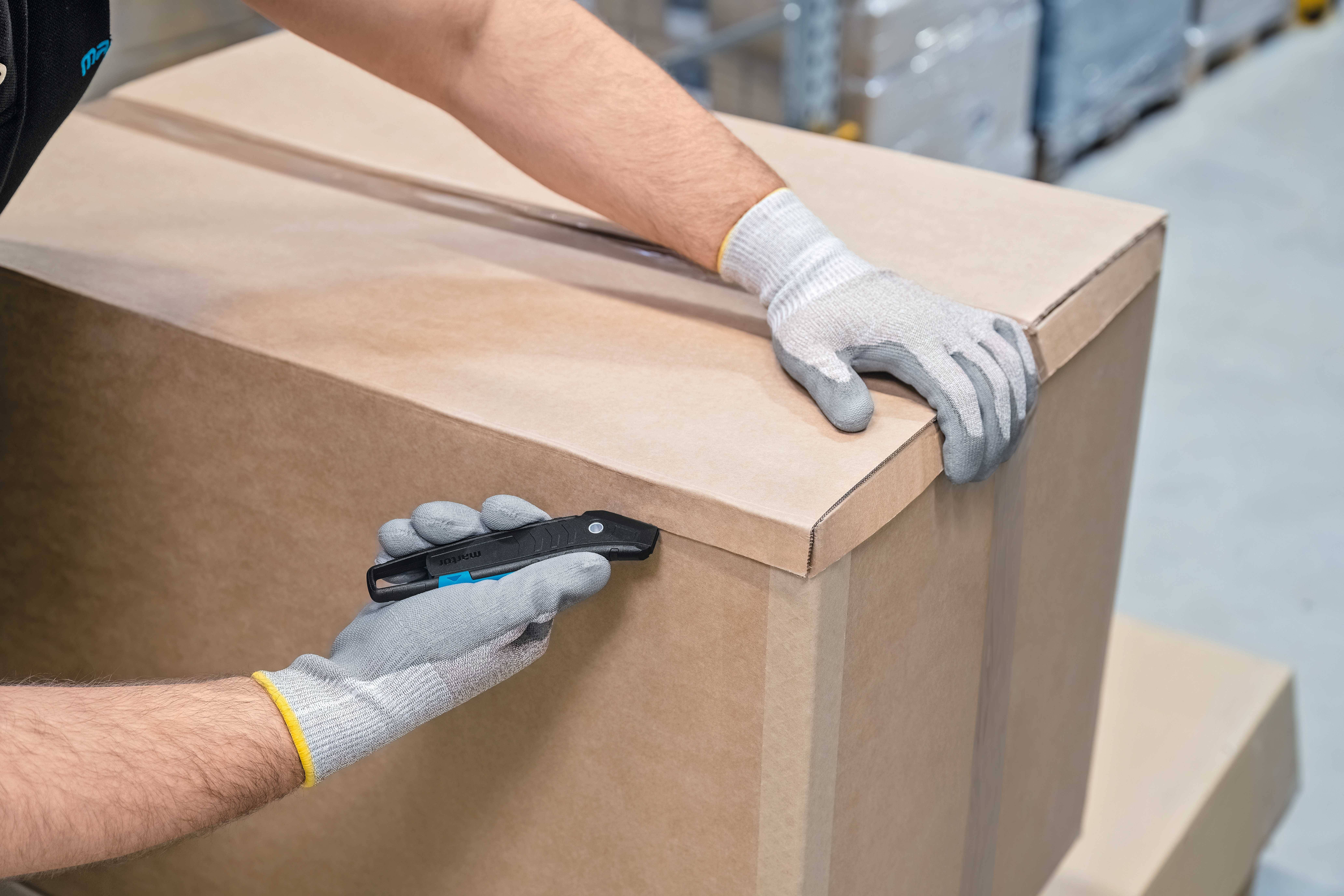 Person in Handschuhen öffnet einen großen Karton mit einem Sicherheitsmesser in einem Lager.