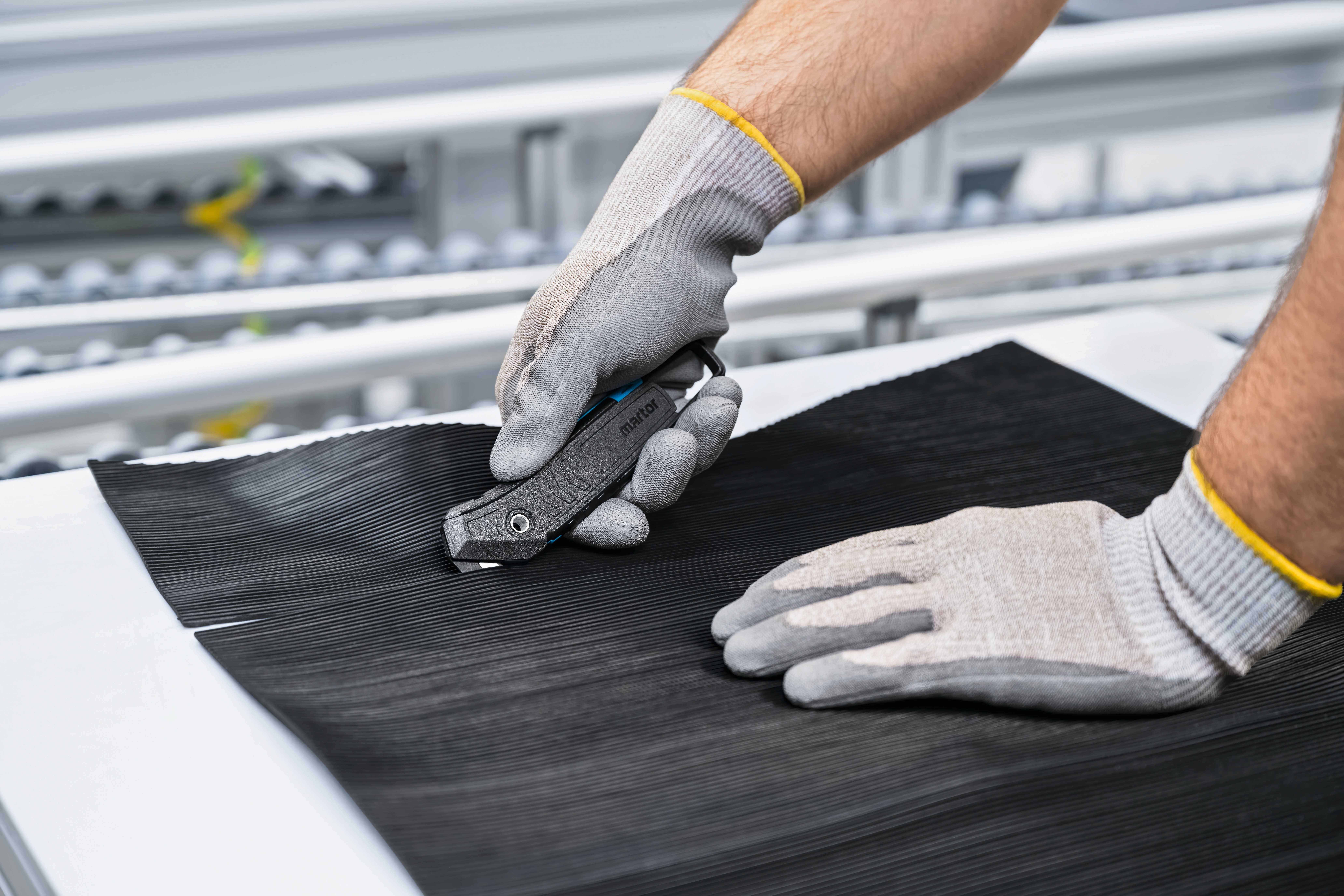 Eine Person schneidet ein schwarzes Material mit einem grauen Messer auf einem Tisch. Sie trägt graue Schutzhandschuhe.