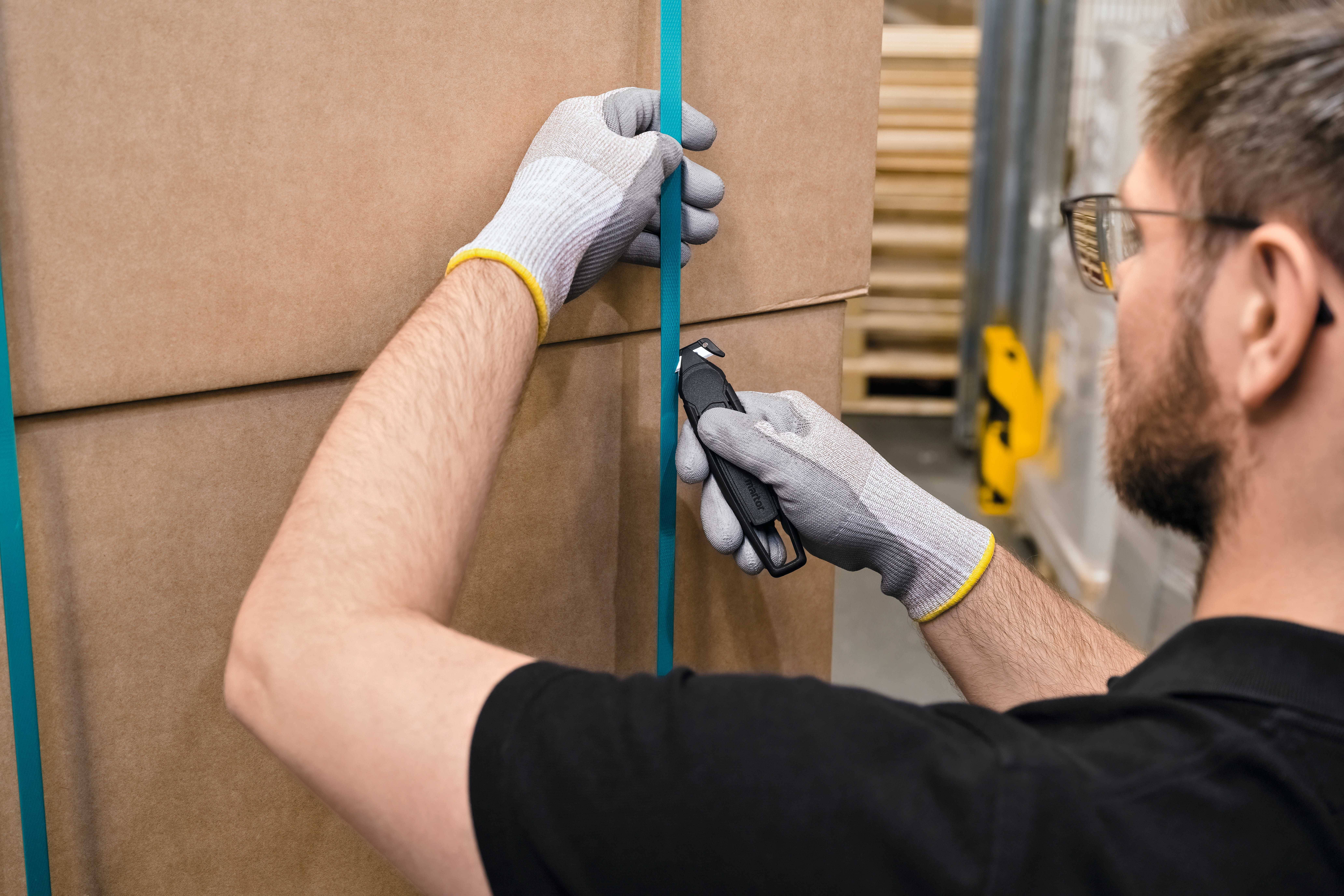 Ein Mann mit Handschuhen schneidet ein Verpackungsband von einem großen Karton. Im Hintergrund sind Regale sichtbar.
