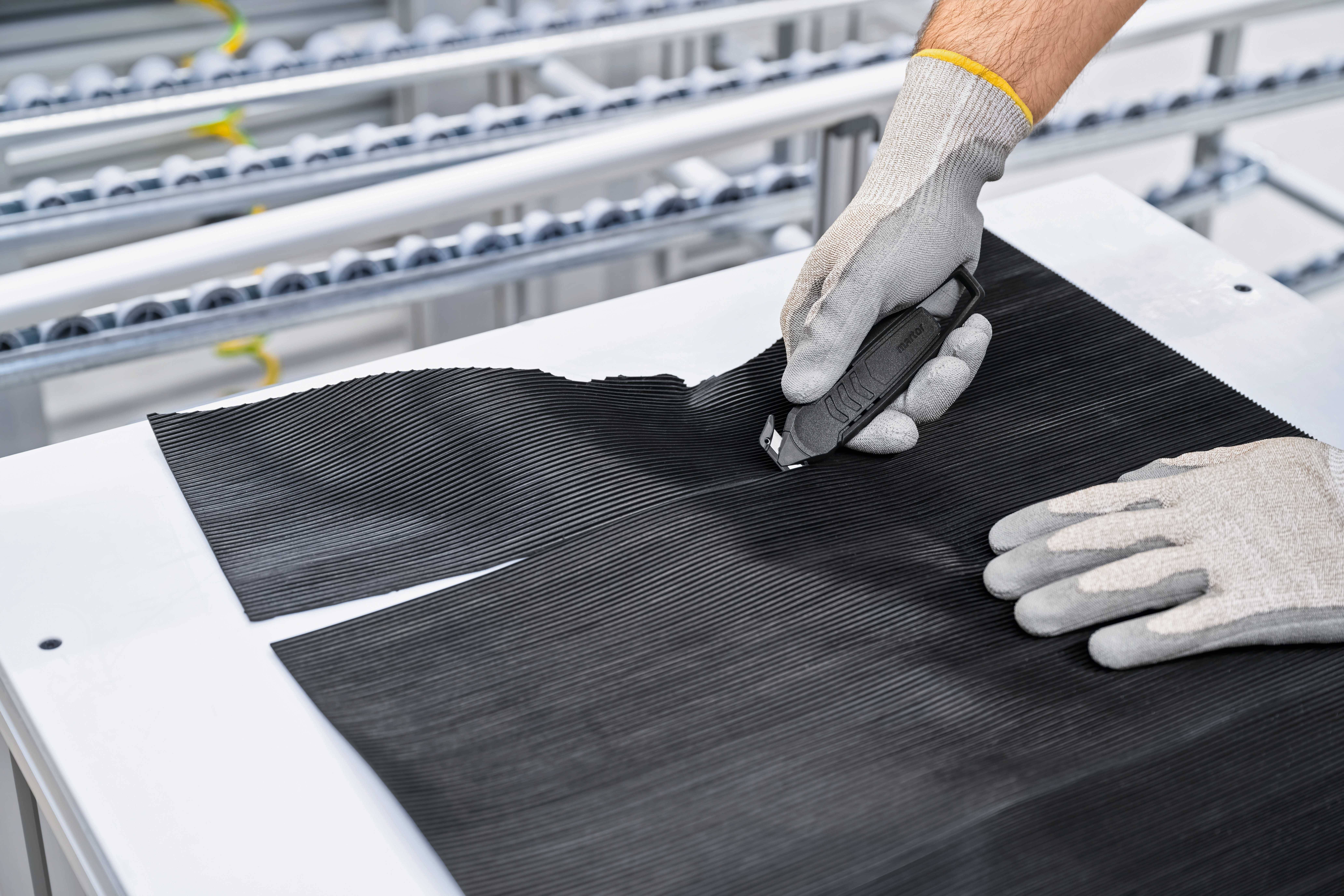 Eine Person schneidet mit einem Messer ein schwarzes Materialstück auf einem weißen Tisch. Sie trägt Handschuhe. Im Hintergrund sind Maschinelemente sichtbar.