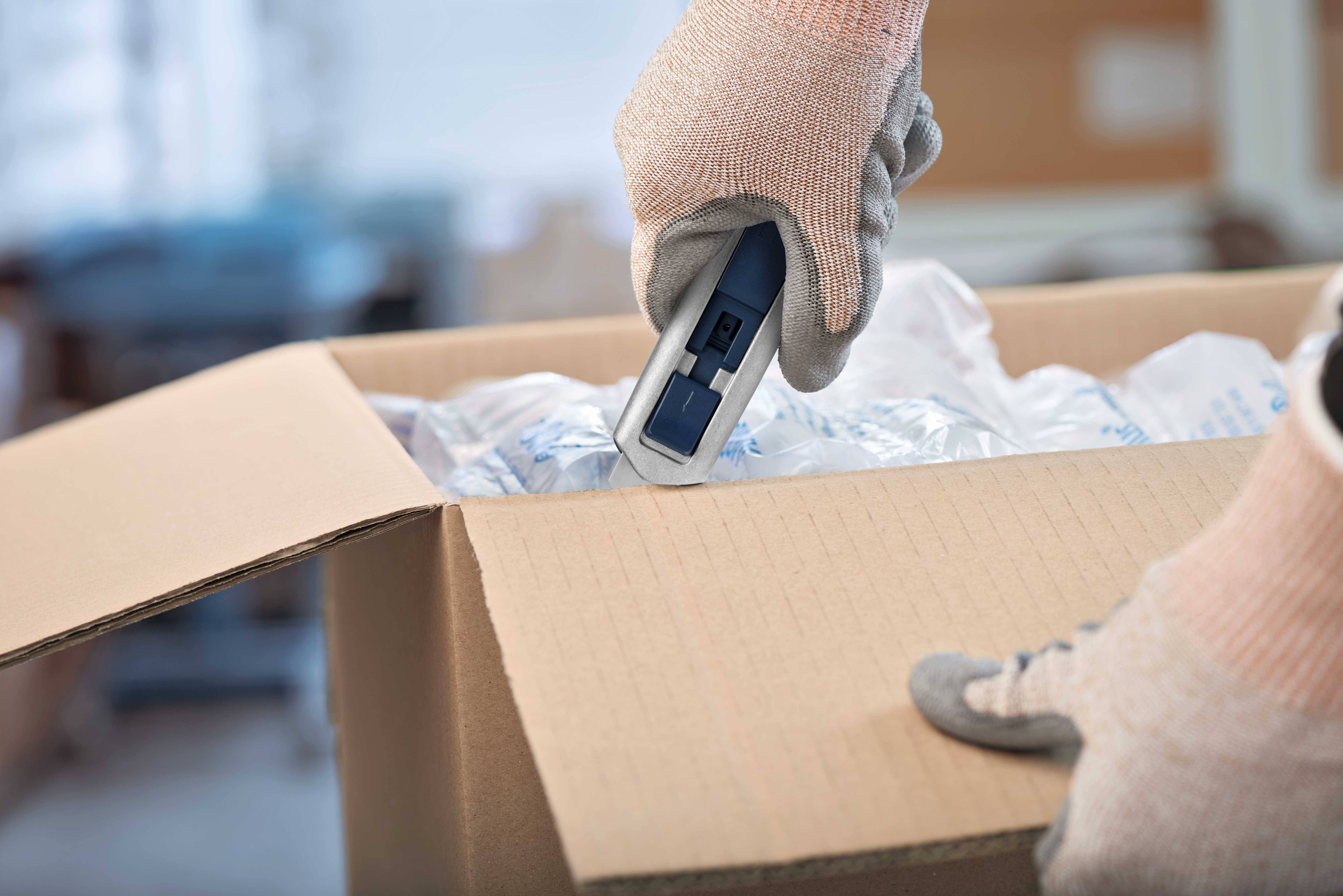 Eine Person mit Handschuhen öffnet mit einem Kartonschneider einen braunen Karton, in dem sich Luftpolsterfolie befindet.