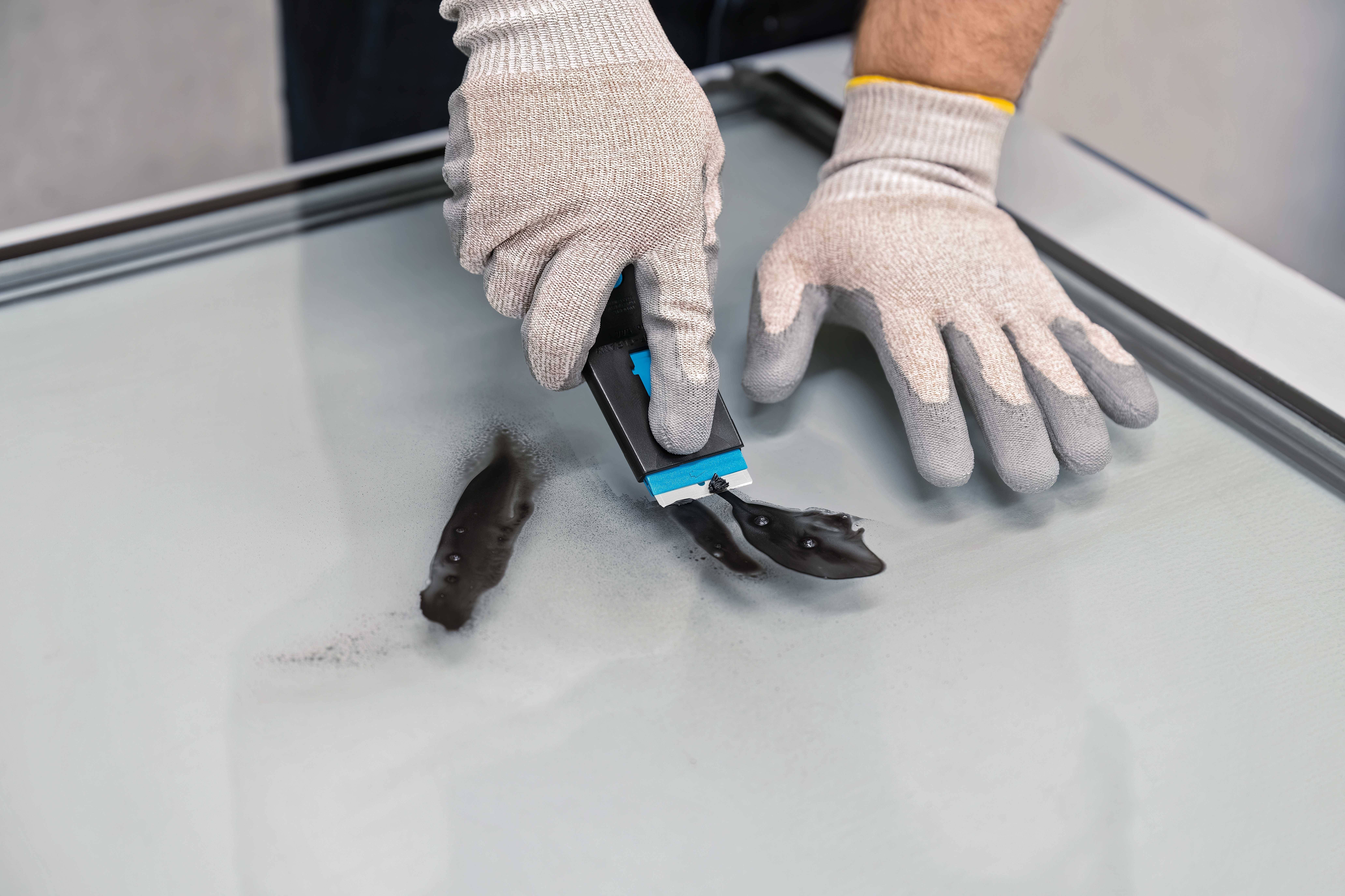 Person trägt Schutzhandschuhe und reinigt eine Glasoberfläche mit einem Schaber.
