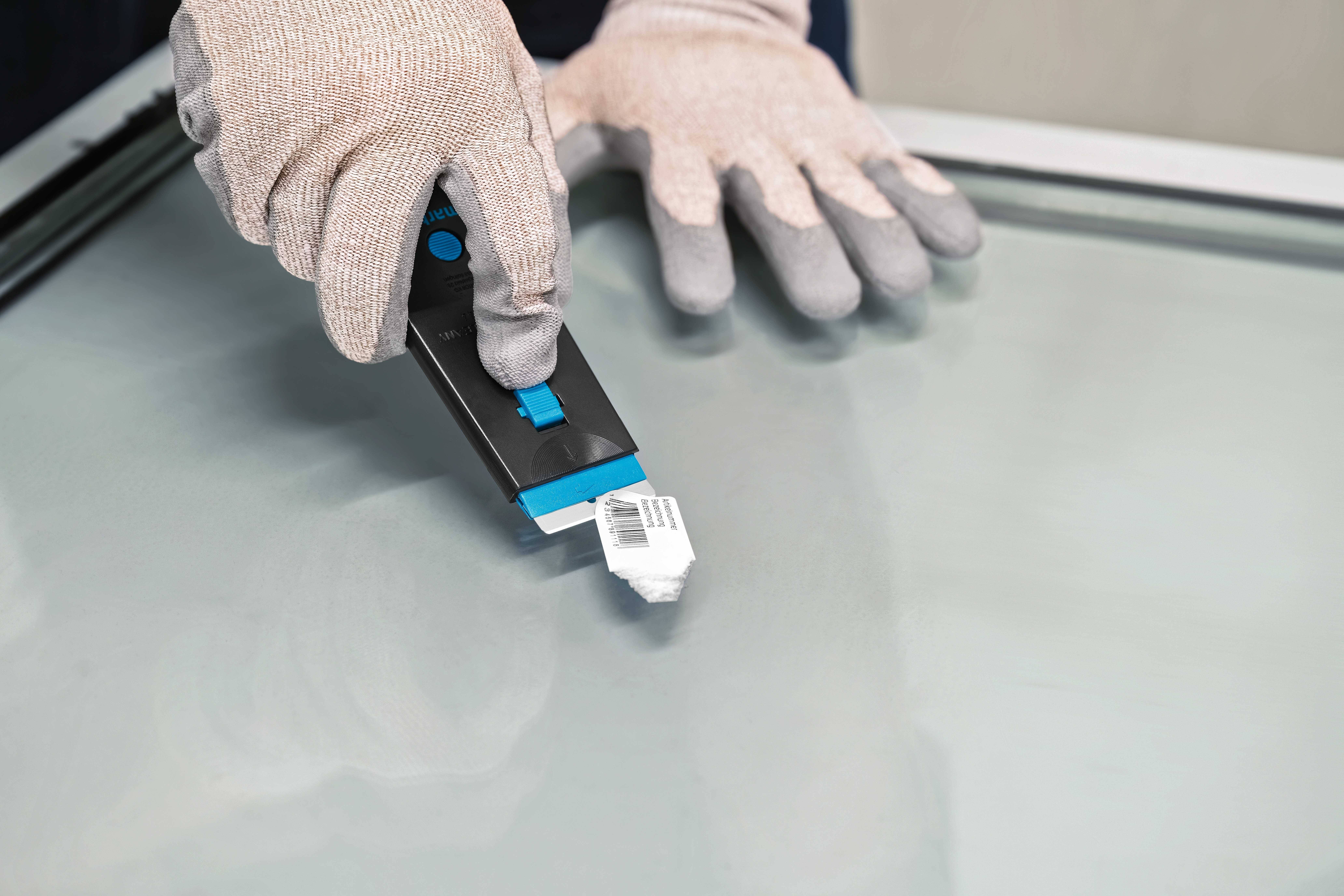 Eine Person entfernt einen Barcode-Aufkleber von einer Glasscheibe mit einem Schaber, trägt dabei graue Handschuhe.