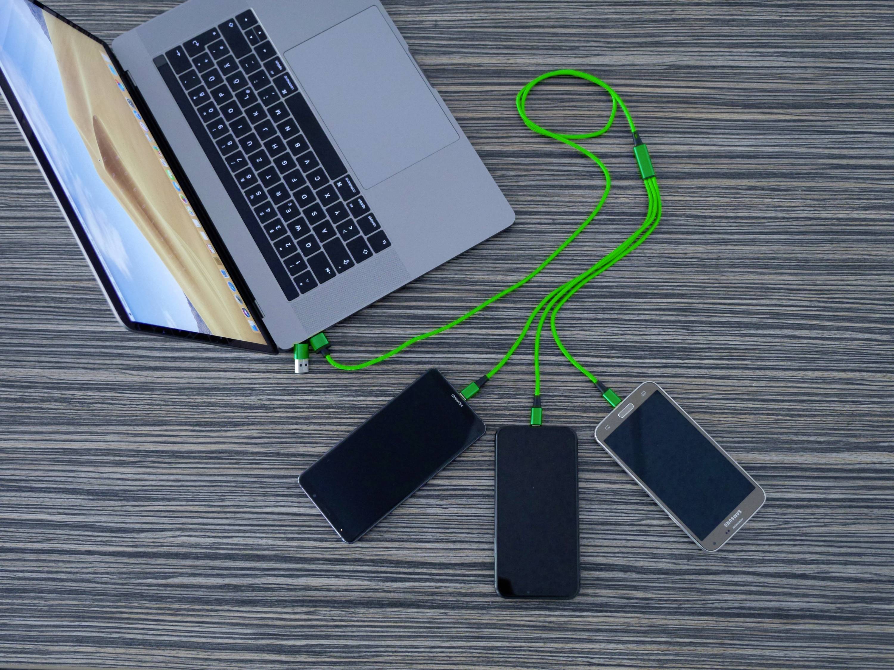 Ein Laptop mit grünem Kabel, das drei Smartphones auflädt. Alle Geräte liegen auf einem Holztisch.