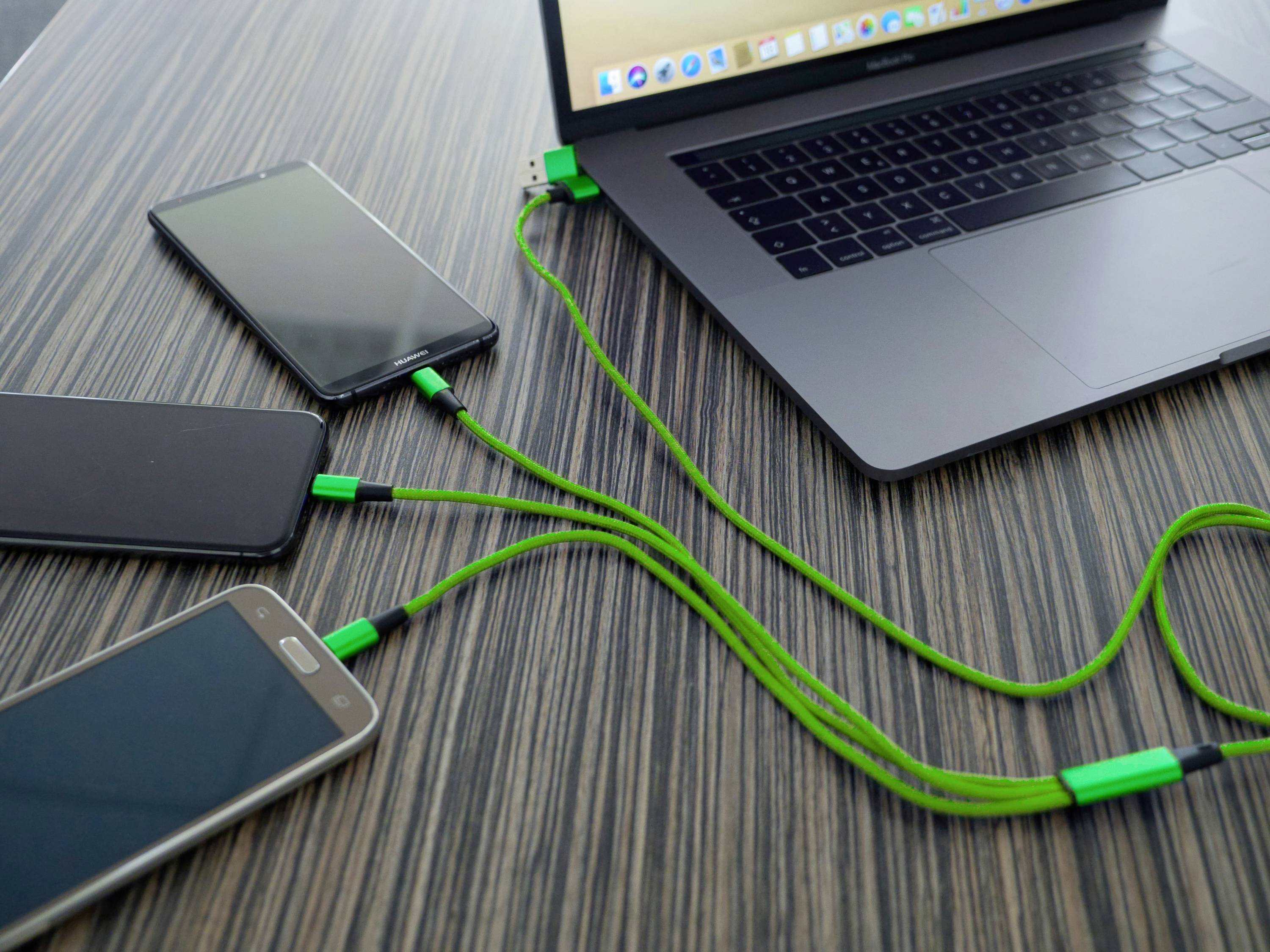 Ein Laptop lädt gleichzeitig drei Smartphones über grüne USB-Kabel auf, die auf einem Holztisch liegen.
