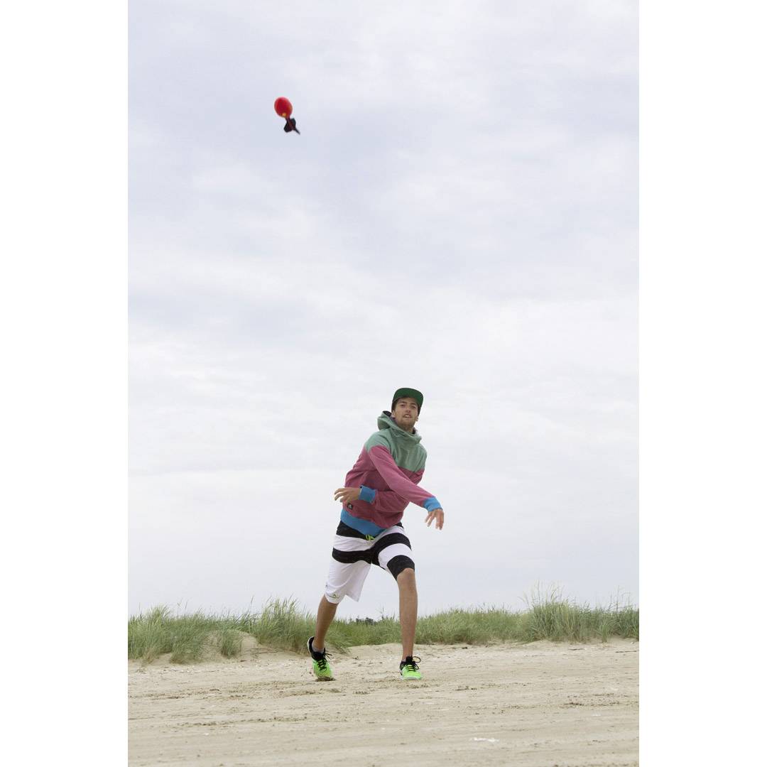Eine Person am Strand wirft einen roten Pokéball in die Luft. Im Hintergrund ist ein bewölkter Himmel zu sehen.