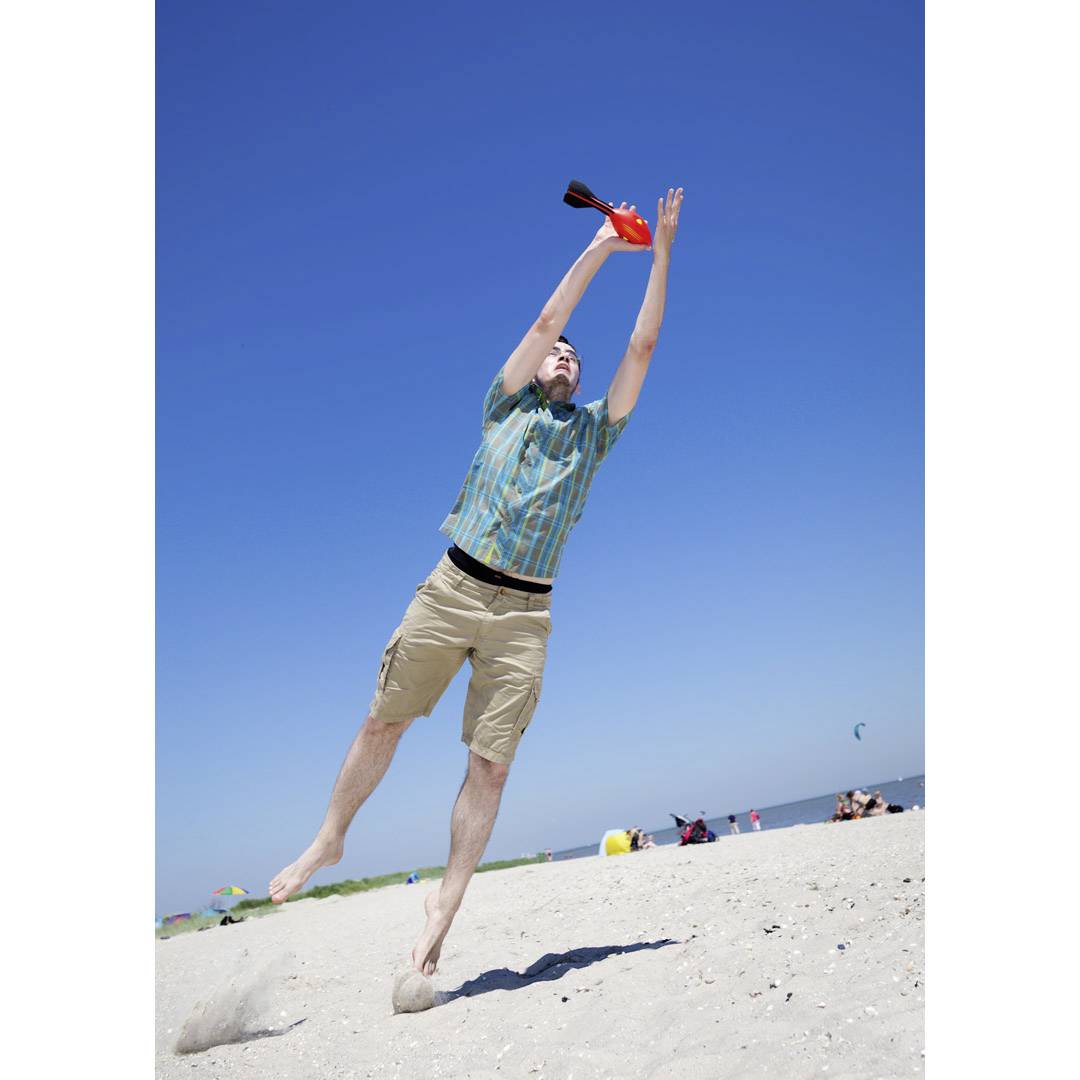 Ein Mann springt am Strand und fängt ein orangefarbenes Objekt. Im Hintergrund ist der blaue Himmel und Meer zu sehen.