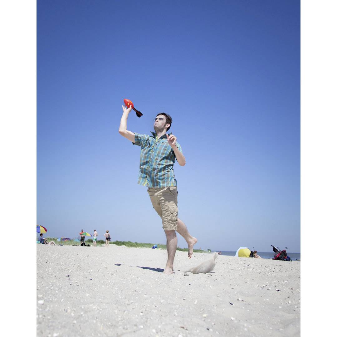 Ein Mann wirft einen Frisbee am Sandstrand. Der Himmel ist klar und blau, mehrere Personen sind im Hintergrund.