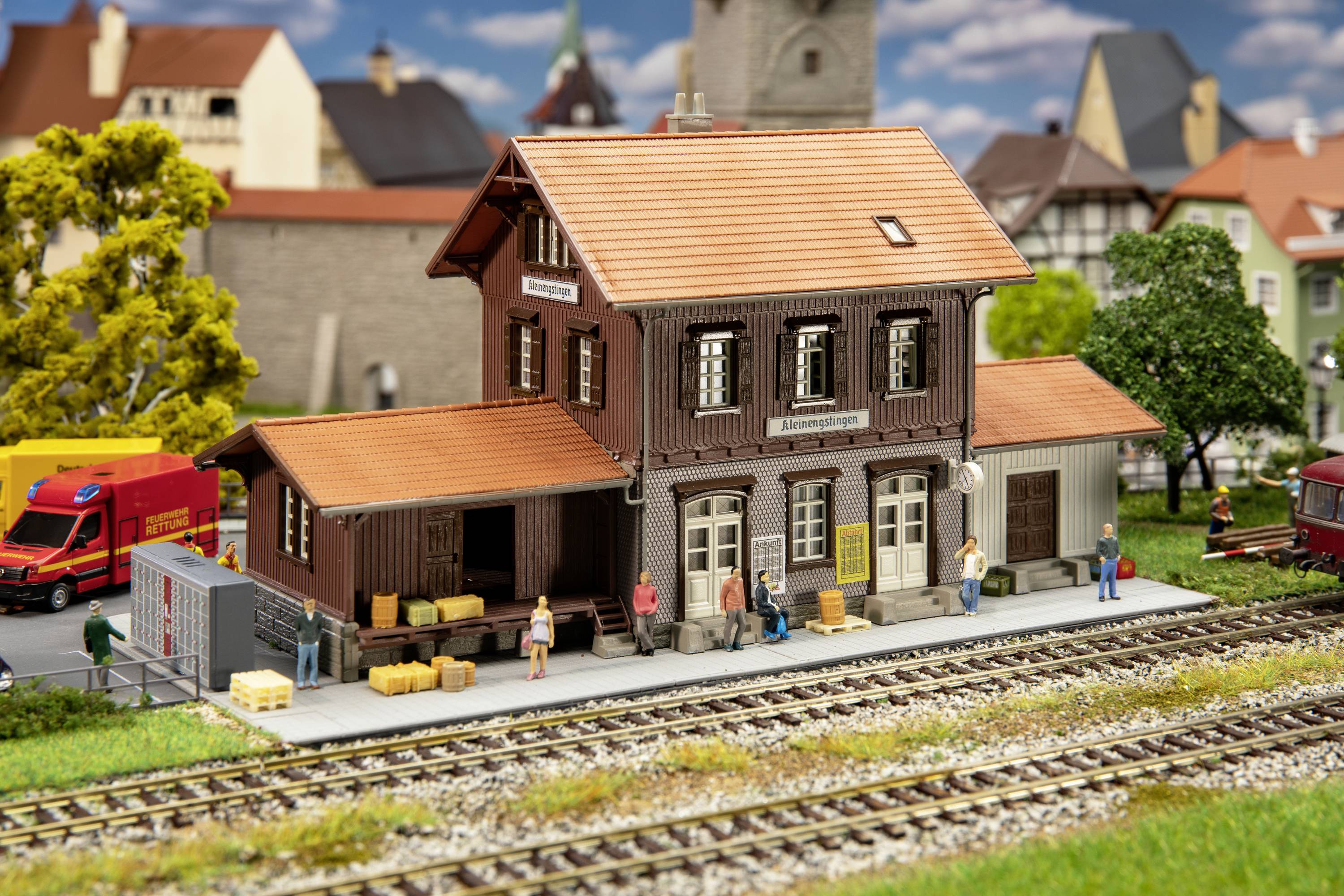 Ein Modellbahnhof in ländlicher Umgebung mit Figuren, Lastwagen und Schienen. Der Bahnhof hat ein zweistöckiges Gebäude mit Holzwänden.