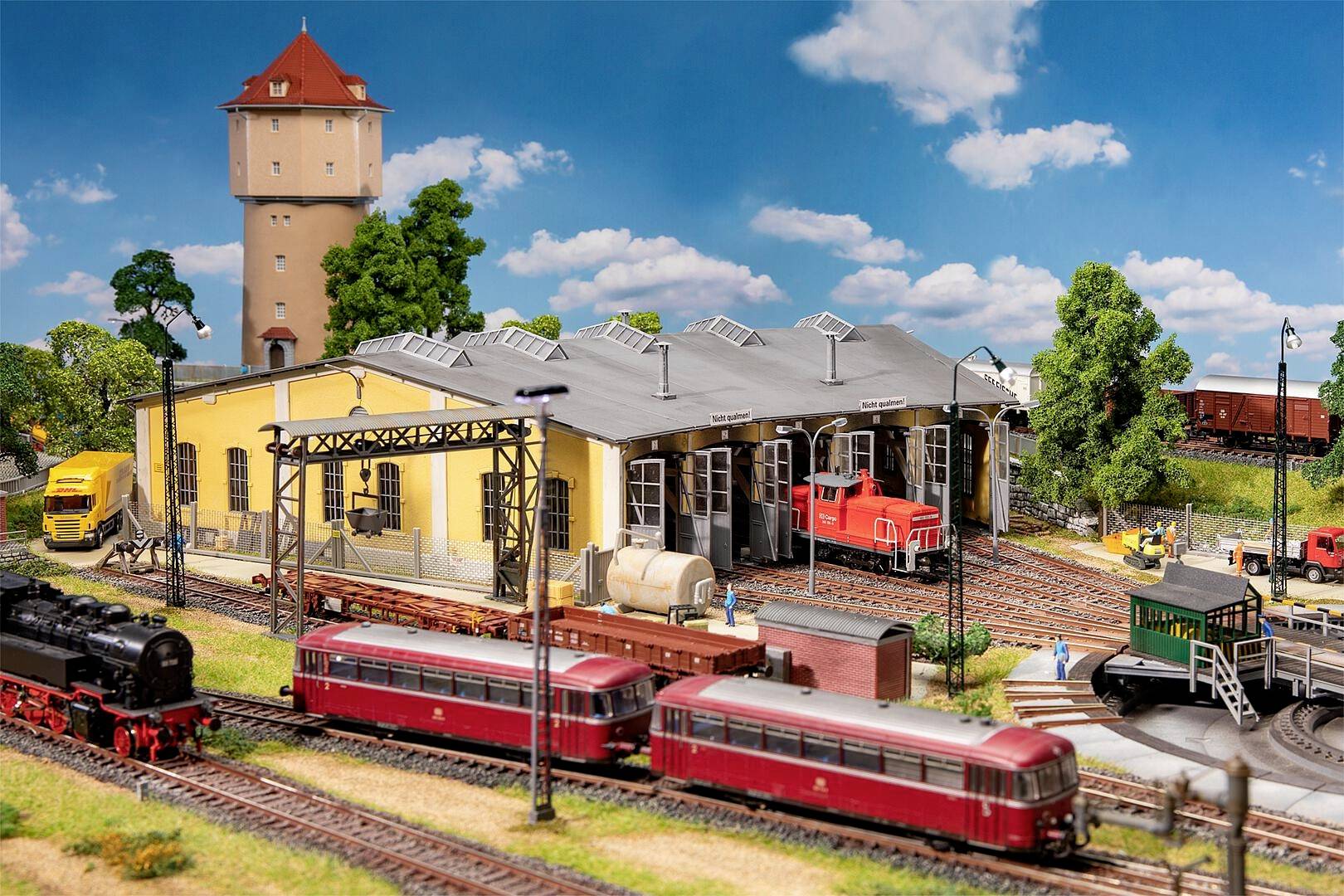 Modelleisenbahnszene mit einer Eisenbahnwerkstatt, mehreren Zügen auf den Gleisen, Bäumen und einem Wasserturm bei klarem Himmel.