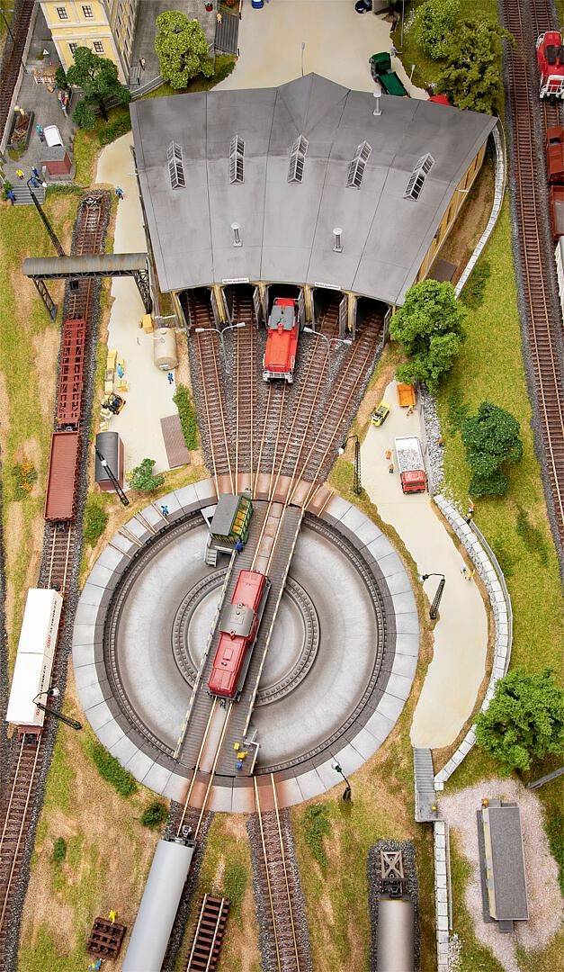 Ein Modellbahndepot mit Lokomotiven auf Gleisen, Drehscheibe in der Mitte, umgeben von Bäumen und weiteren Modellgebäuden.