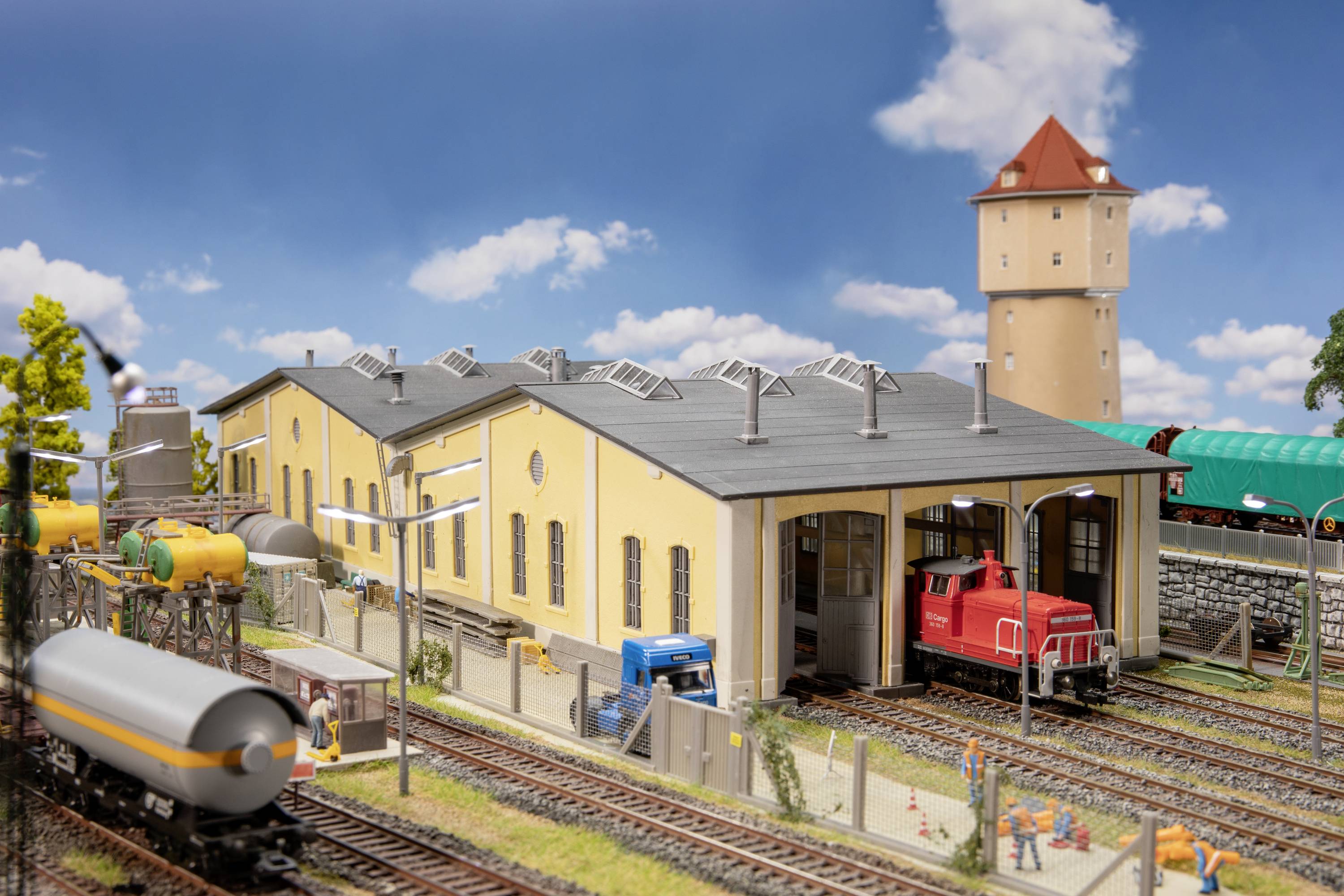Ein Modelleisenbahn-Layout zeigt ein Industriegebiet mit gelbem Fabrikgebäude, einem roten Zug, mehreren Gleisen und einem Wasserturm.