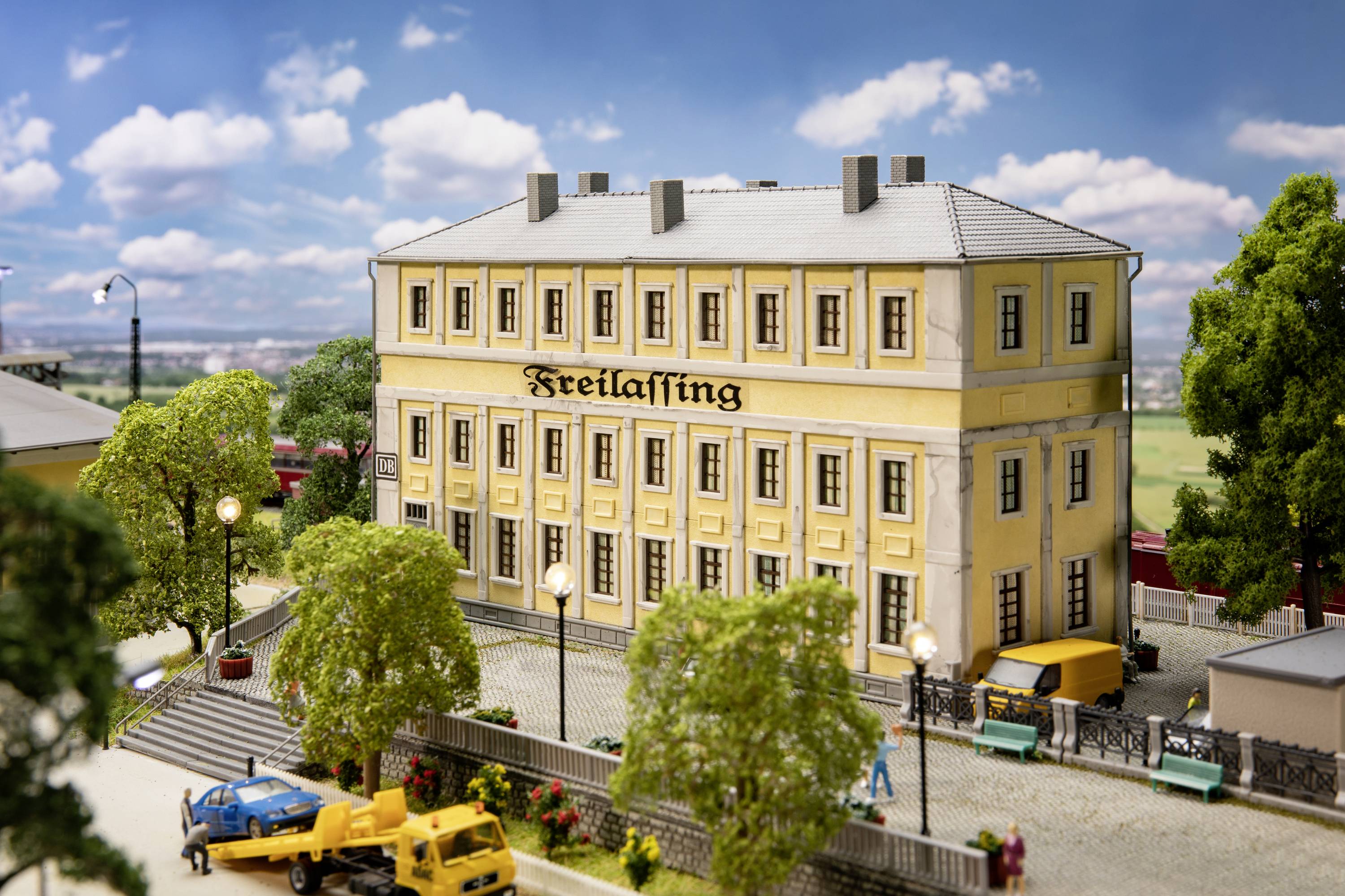 Miniaturdarstellung eines Gebäudes mit der Aufschrift 'Freilassing'. Umgeben von Bäumen und Autos; Modellbahn im Hintergrund.