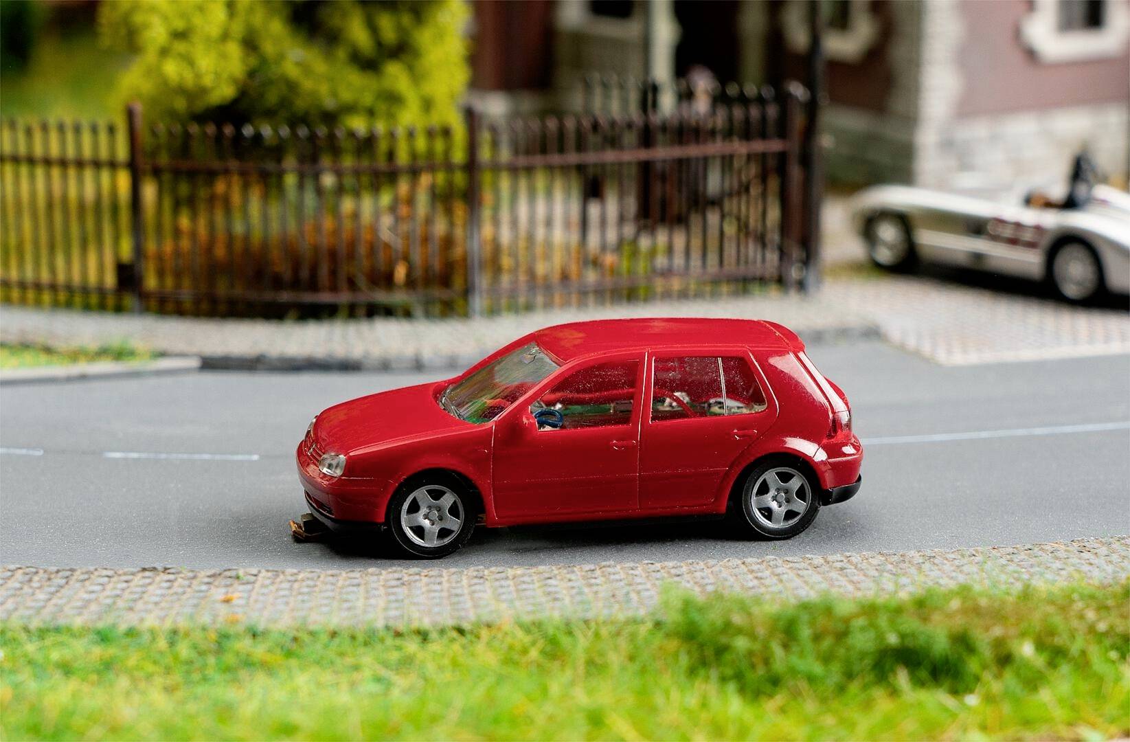 Ein rotes Modellauto steht auf einem grauen Straßendiorama vor einem metallischen Zaun und grünen Pflanzen im Hintergrund.