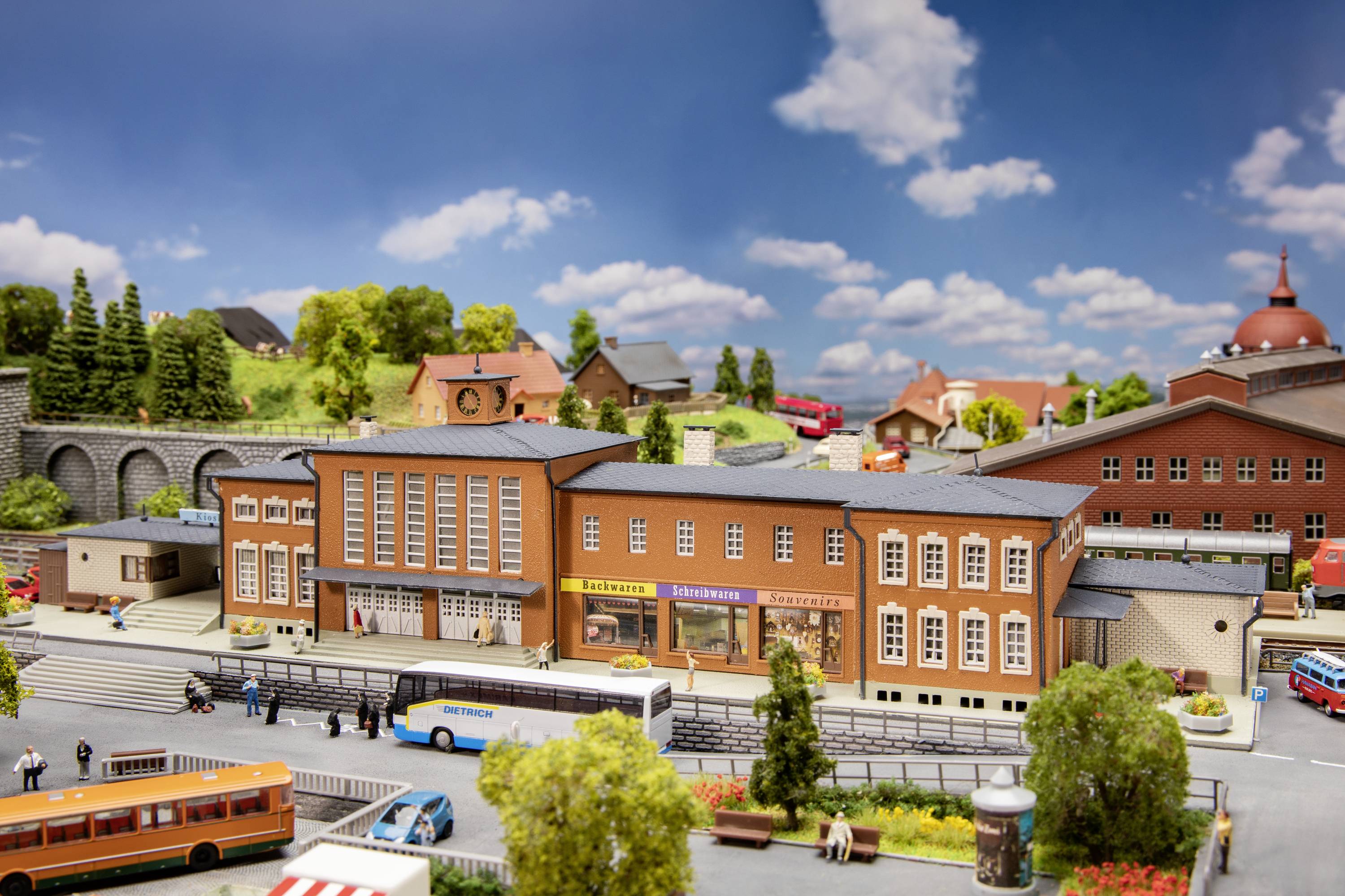 Modellbahn-Szene mit Bahnhof, Bussen und Gebäuden, umgeben von Bäumen und Hügeln, alles unter einem blauen Himmel mit Wolken.