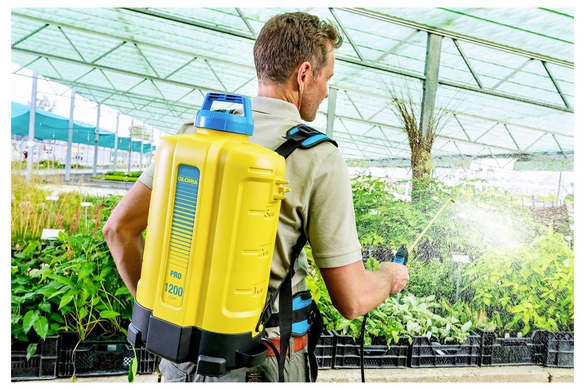 Eine Person mit einem gelben rucksackartigen Sprühgerät bewässert Pflanzen in einem großen Gewächshaus, das mit verschiedenen Grünpflanzen gefüllt ist.