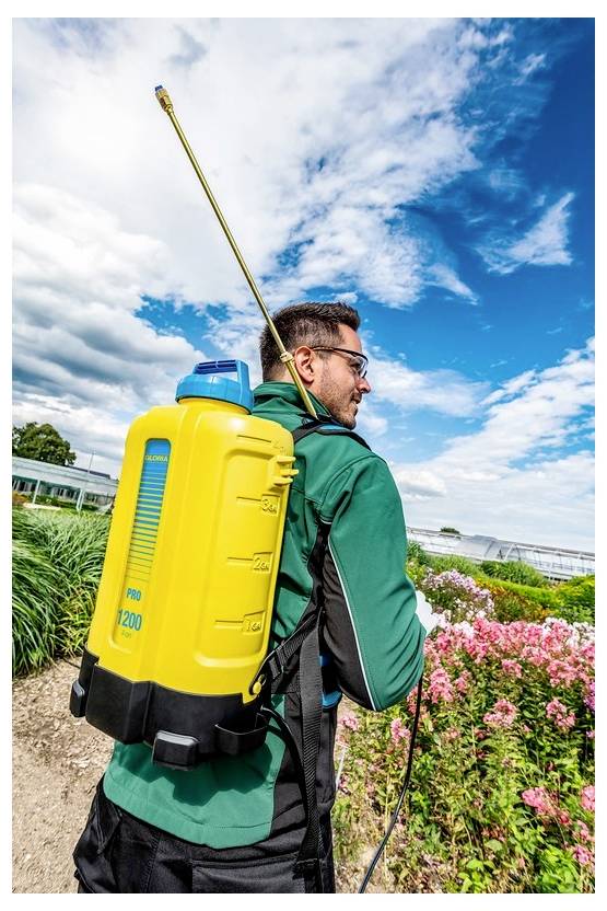 Eine Person in grüner Kleidung trägt einen gelben Rucksack-Sprüher, während sie auf einem Gartenpfad mit blühenden Pflanzen unter blauem Himmel spaziert.