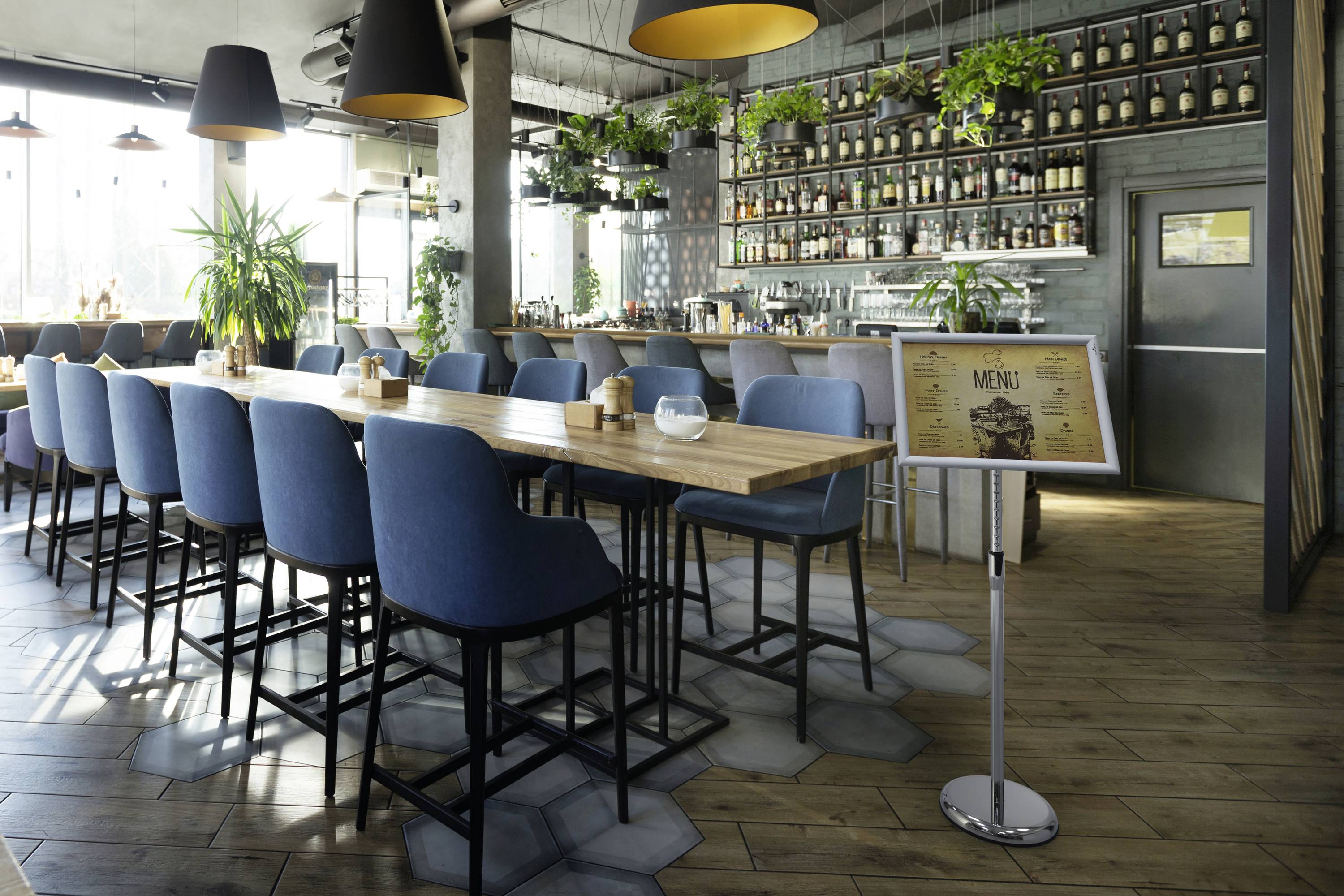 Ein modernes Café mit einer langen Holztheke, blauen Stühlen und Pflanzen. Eine Menütafel steht rechts vom Tresen.