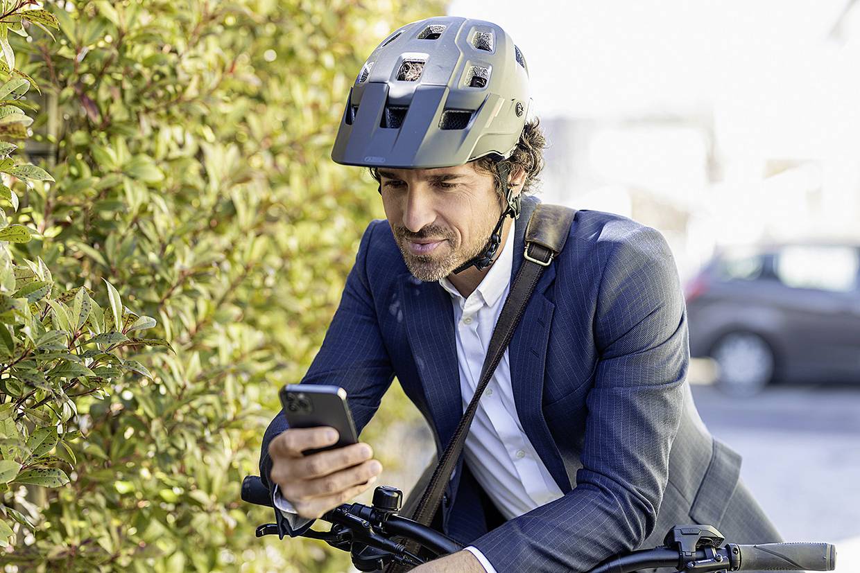 Ein Mann im Anzug, trägt einen Helm und hält ein Smartphone, während er auf einem Fahrrad sitzt, neben einer Hecke auf einer Straße.
