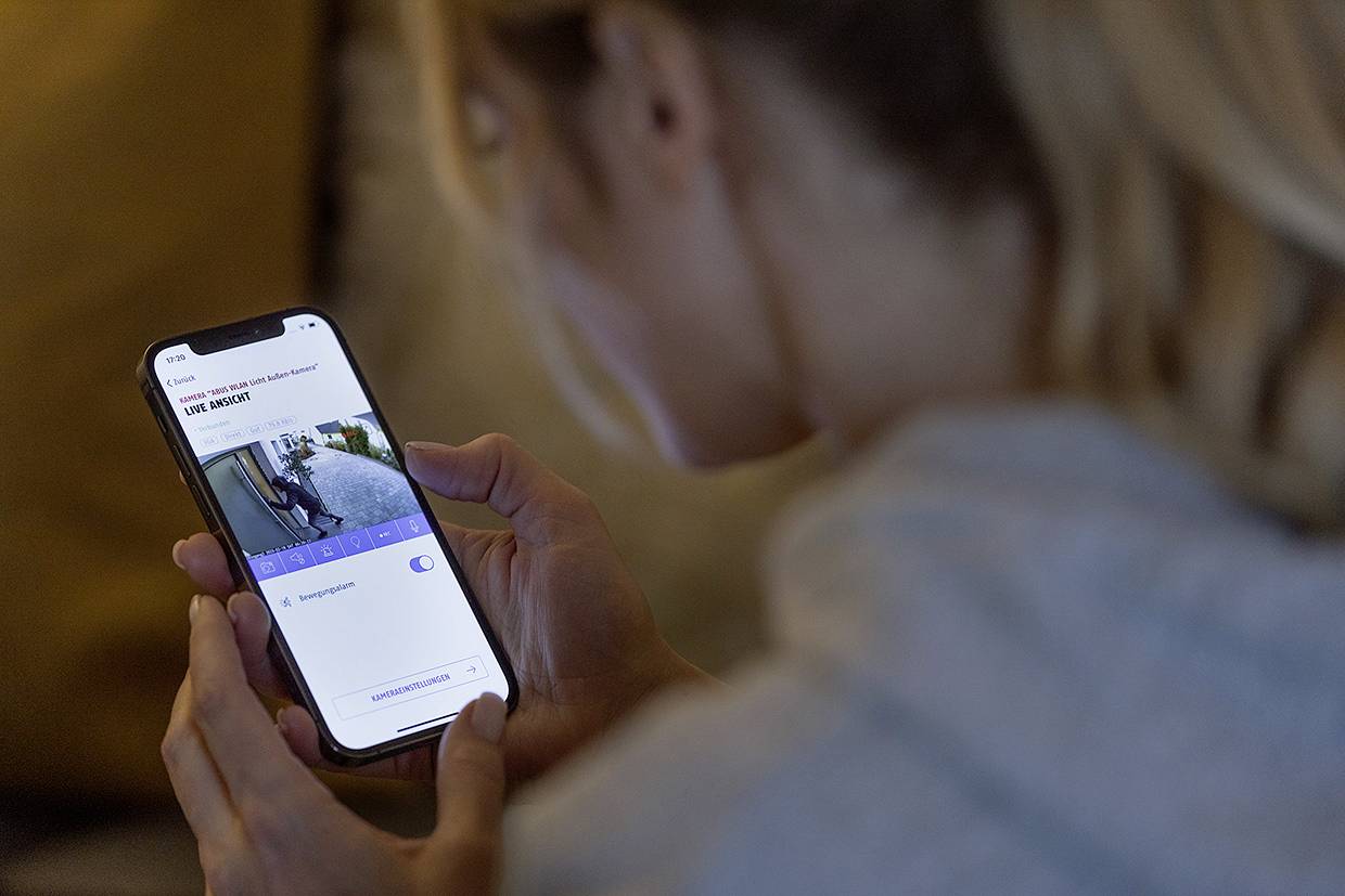 Eine Person betrachtet ein Smartphone, das eine Live-Überwachungskamera zeigt. Die Kamera zeigt den Eingang eines Hauses.