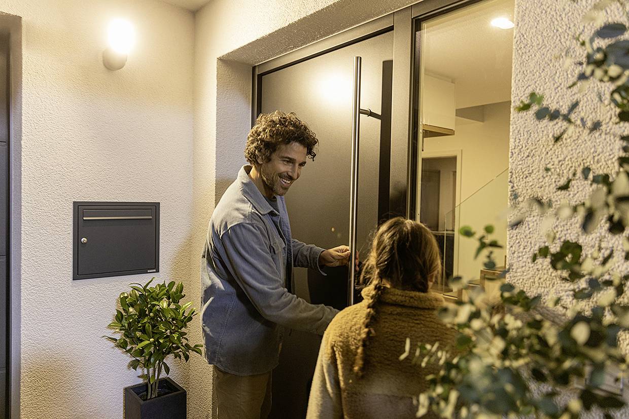 Ein Mann öffnet lächelnd die Haustür für eine Person mit langen Haaren. Sie stehen vor einem beleuchteten Eingang mit Pflanzen daneben.