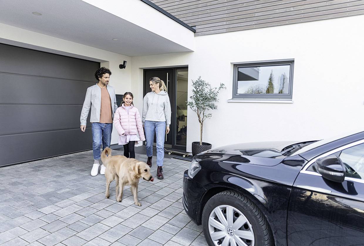 Eine Familie mit einem Hund geht lächelnd auf einem gepflasterten Weg vor einem modernen Haus. Ein schwarzes Auto ist im Vordergrund geparkt.
