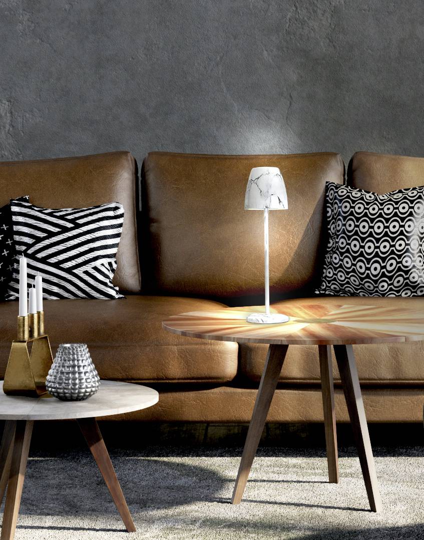 Ein Wohnzimmer mit braunem Ledersofa, zwei gemusterten Kissen, zwei runden Holztischen und einer kleinen Lampe auf dem Tisch.