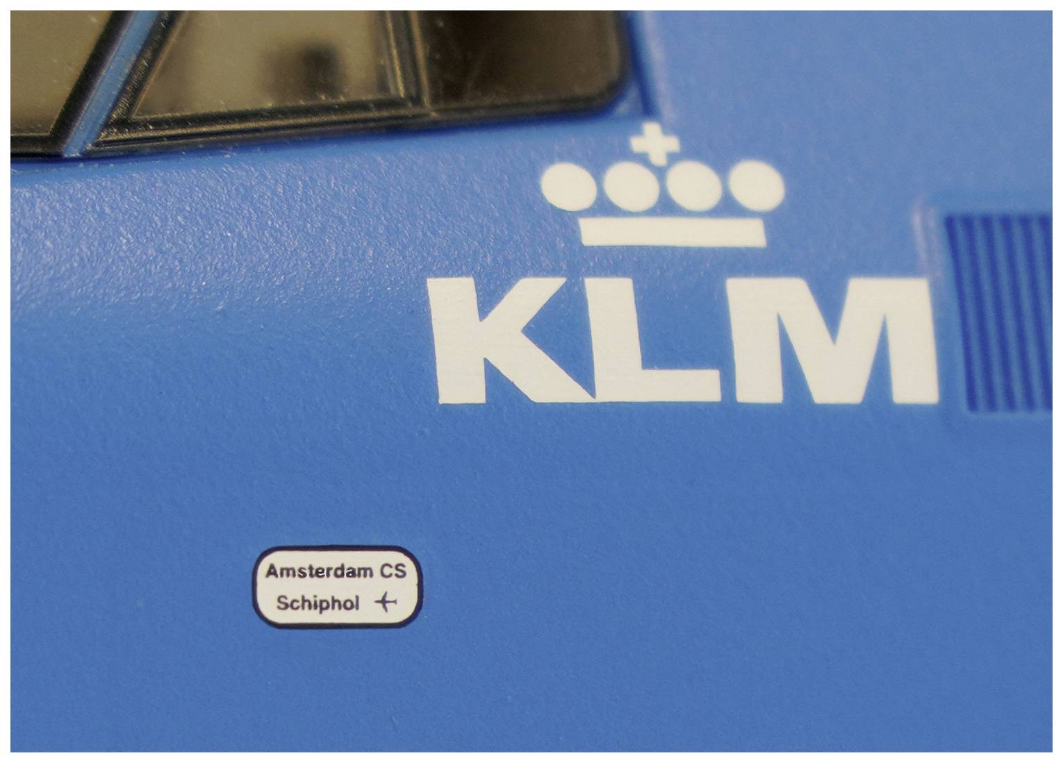 Blauer Zug mit KLM-Logo, angedeuteten Zügen des niederländischen Eisenbahnnetzes, darunter 'Amsterdam CS' und 'Schiphol'.