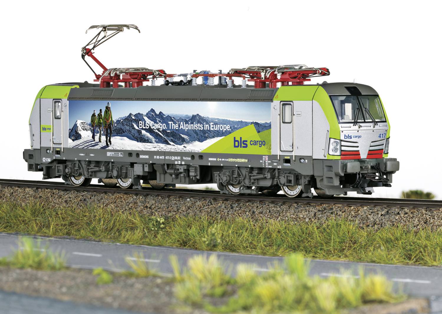 Eine grüne Lokomotive mit Werbung 'bls cargo, The Alpinists in Europe', fährt auf Bahngleisen. Hintergrund zeigt Berglandschaft.