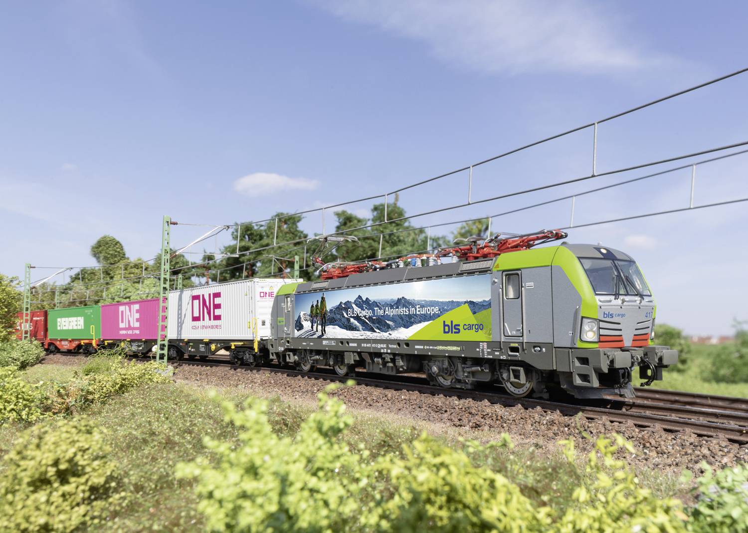 Ein Güterzug fährt tagsüber auf einer Eisenbahnstrecke. Der Zug transportiert Container mit Werbeaufdrucken. Im Hintergrund sind Bäume und Stromleitungen zu sehen.