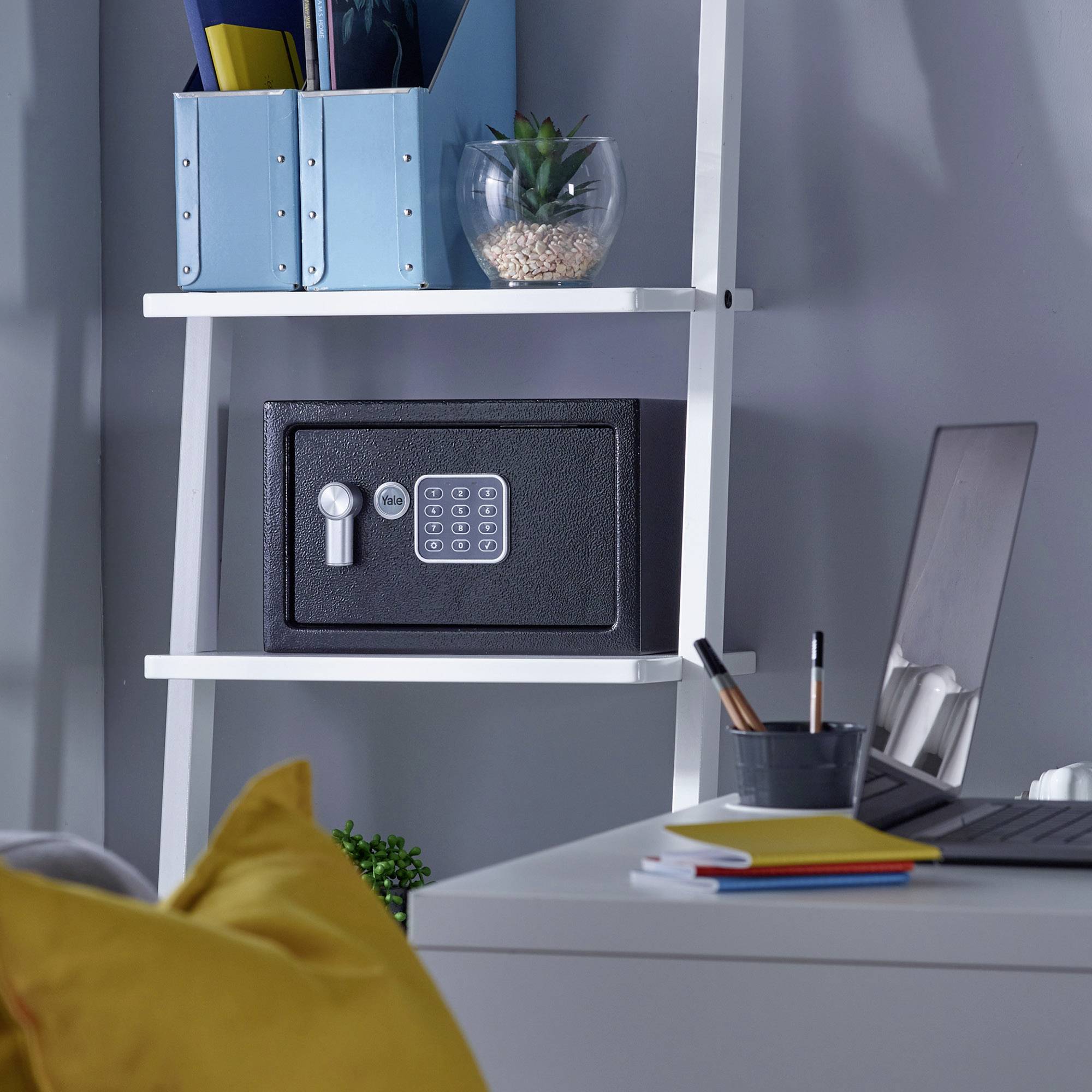 Ein weißes Regal mit einer Safe-Box, Büchern und einem Glas mit Pflanze darauf. Ein Laptop und Notizbuch liegen auf einem Schreibtisch daneben.