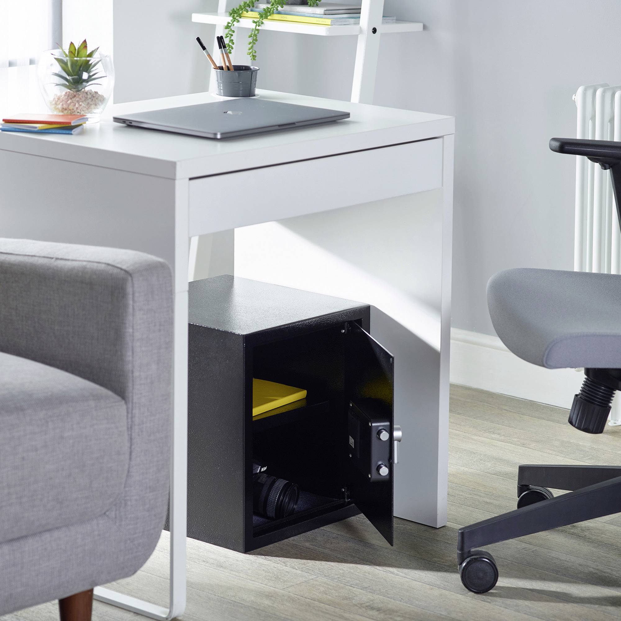 Ein offener Tresor unter einem Schreibtisch zeigt gelbe Dokumente und eine Kamera im Inneren. Ein Laptop und Pflanzen auf dem Tisch.