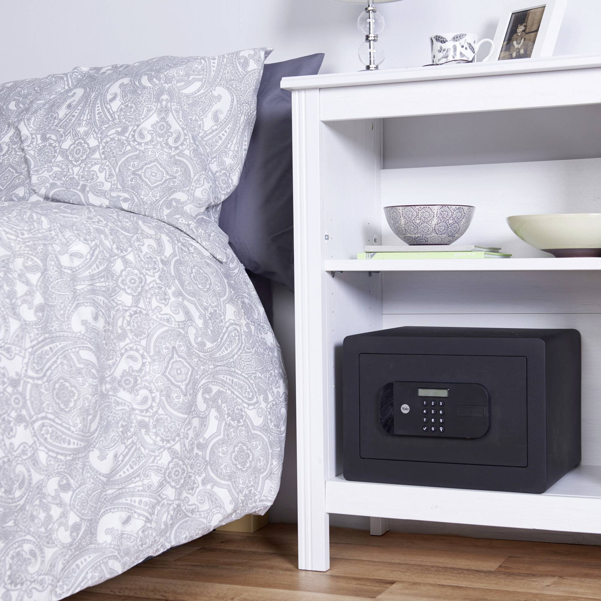 Ein Schlafzimmer mit einem Bett links und einem weißen Nachttisch rechts. Auf dem Tisch steht ein Tresor in einem Regal.