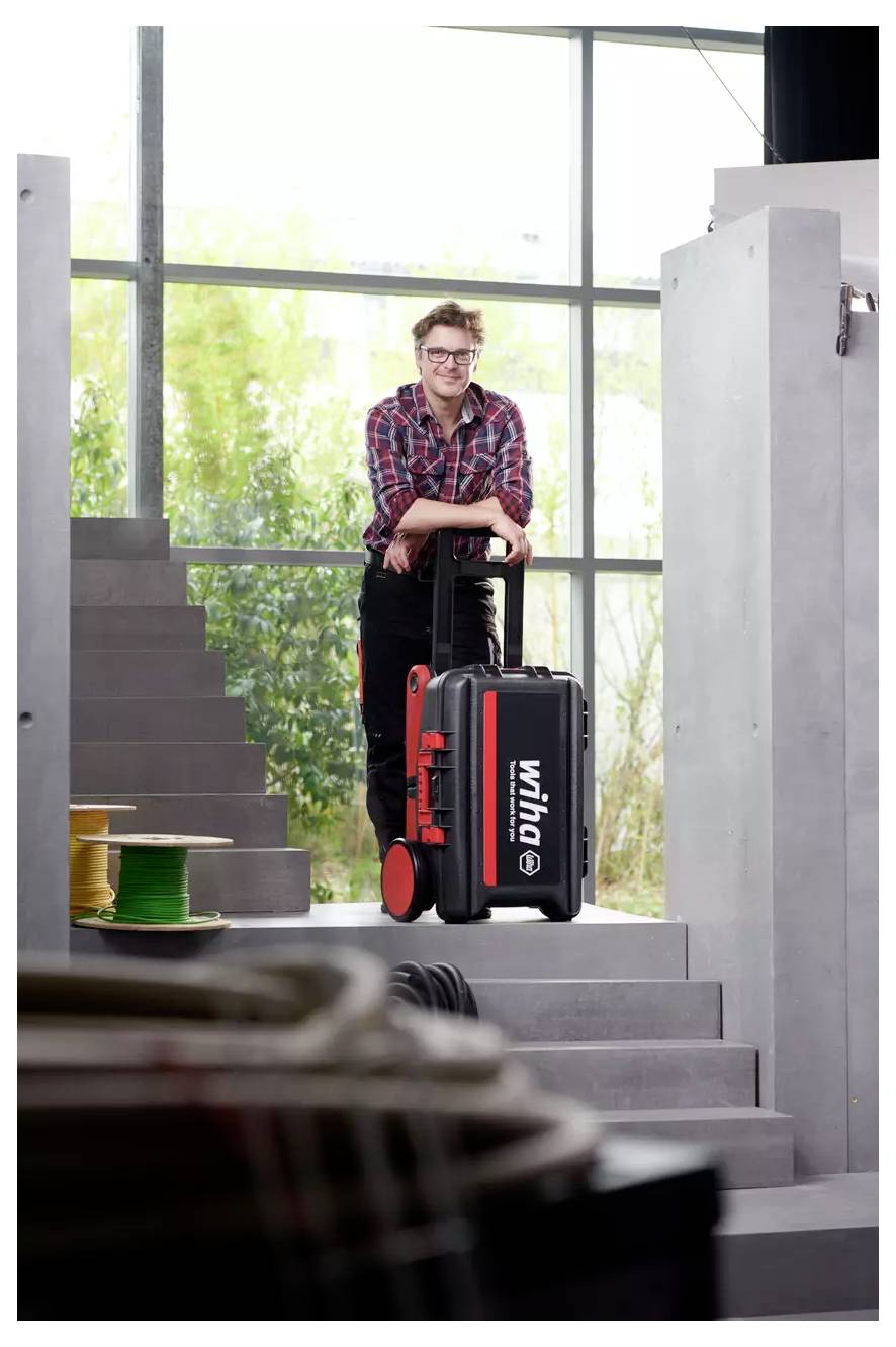 Eine Person in einem karierten Hemd lächelt, während sie sich auf einen Werkzeugkasten mit Rollen lehnt, der mit „wiha