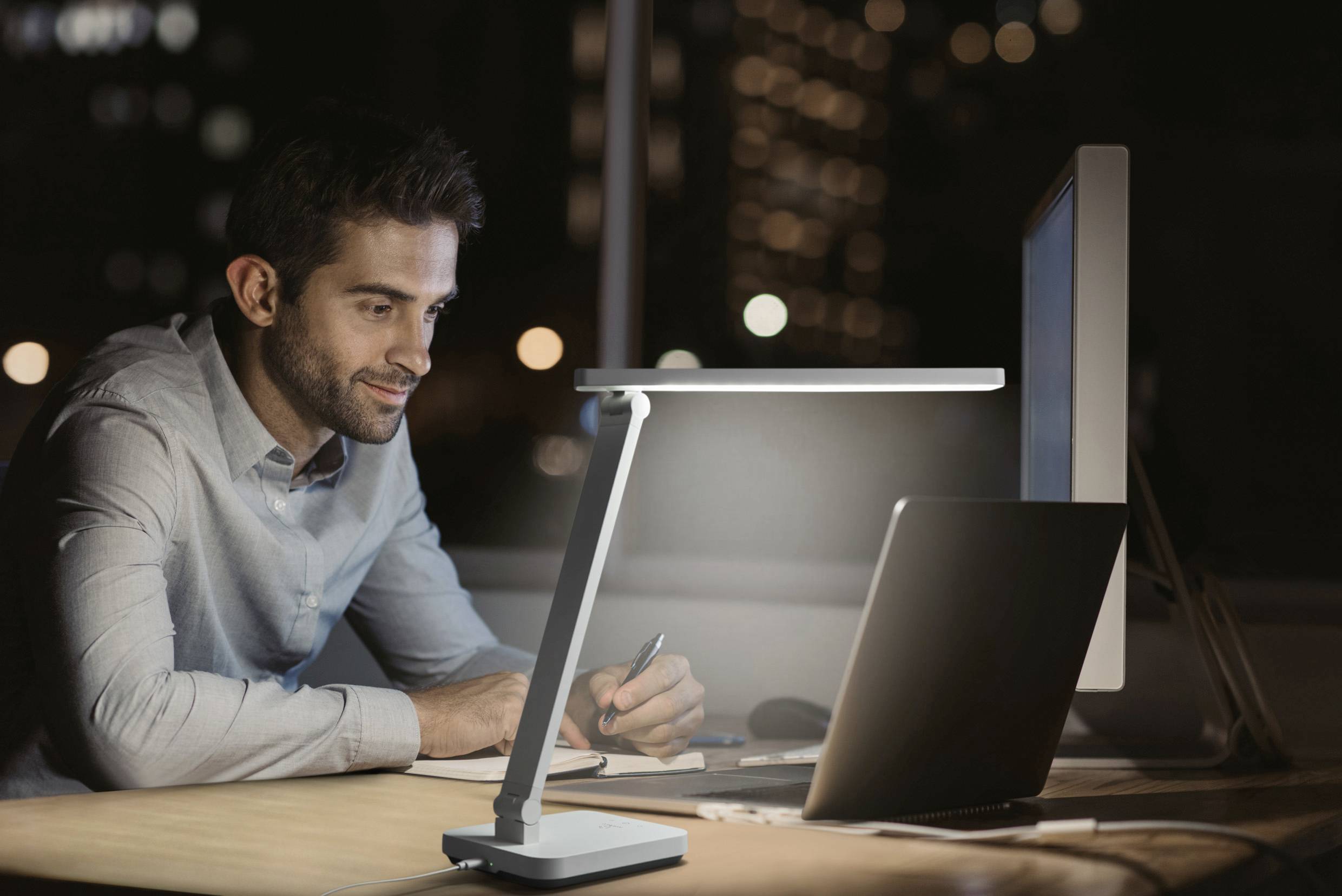 Mann arbeitet abends am Schreibtisch mit Computer und Notizbuch, während eine Tischlampe den Bereich beleuchtet. Bürogebäude im Hintergrund.